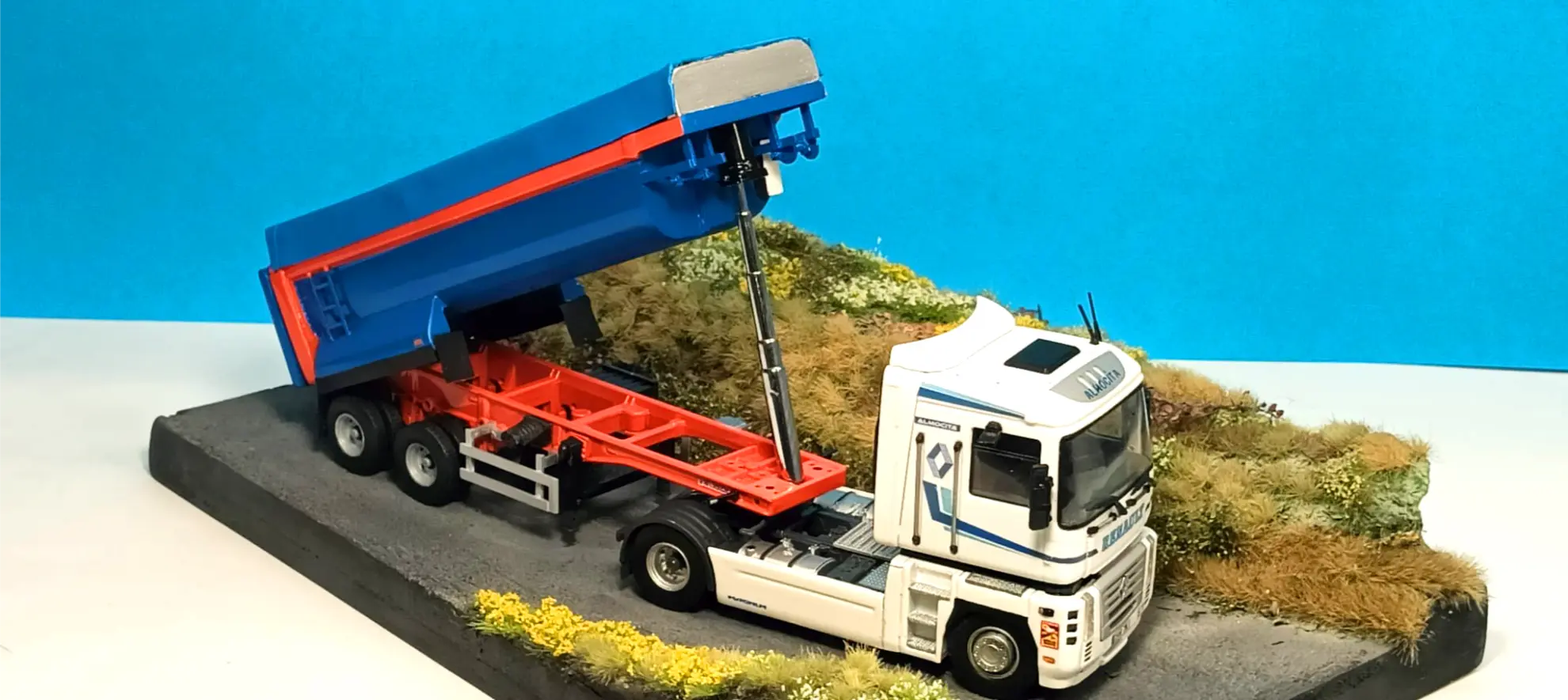 Un modelo de camión volquete blanco y azul de Italeri descarga la carga en una carretera en miniatura con vegetación, sobre un fondo azul: la inspiración perfecta si está aprendiendo cómo hacer una maqueta de camión o explorando los camiones a escala 1:24.