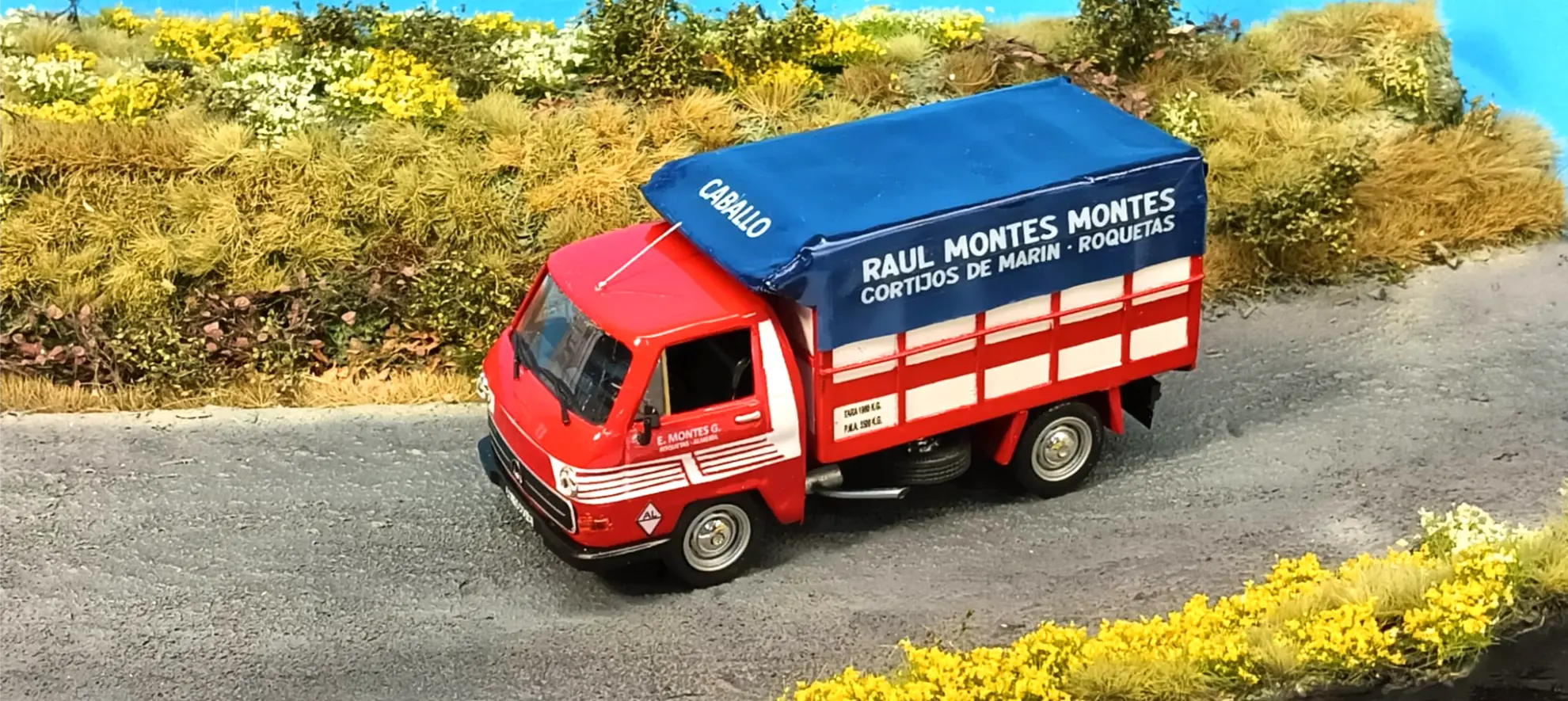 Un camión de reparto rojo con una lona azul circula por un camino rural rodeado de vegetación y flores amarillas, inspiración perfecta para Italeri camiones o ideas sobre cómo hacer una maqueta de camión.