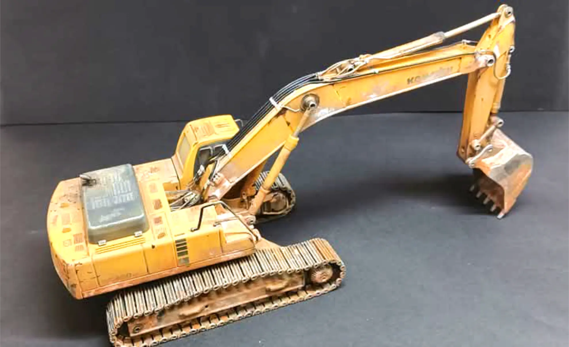 Una excavadora de juguete amarilla con orugas y un largo brazo excavador, perfecta para coleccionismo camiones, mostrada sobre un fondo negro.