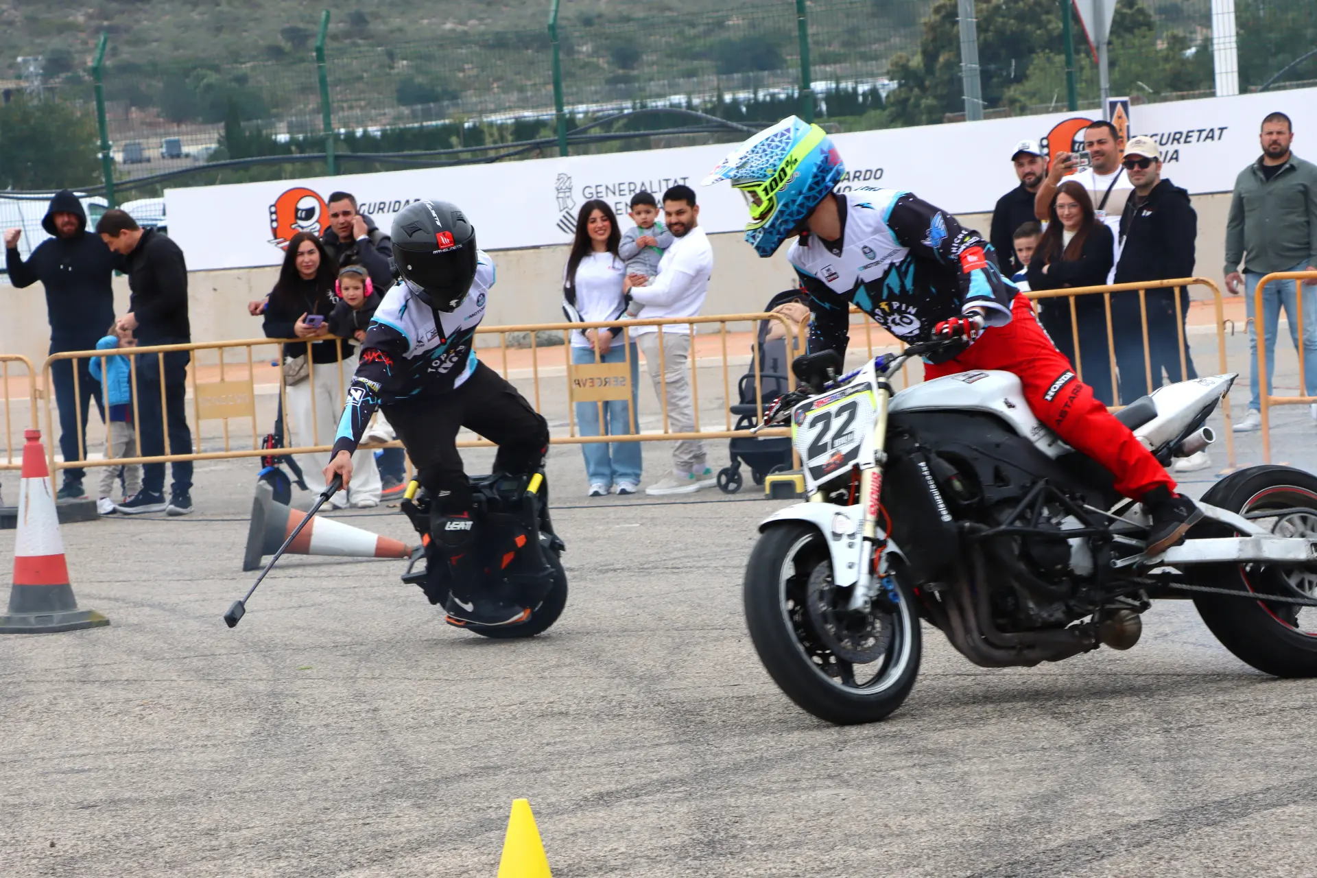 Un motociclista y un monociclista compiten en una prueba al aire libre, observados por personas detrás de unas barreras.