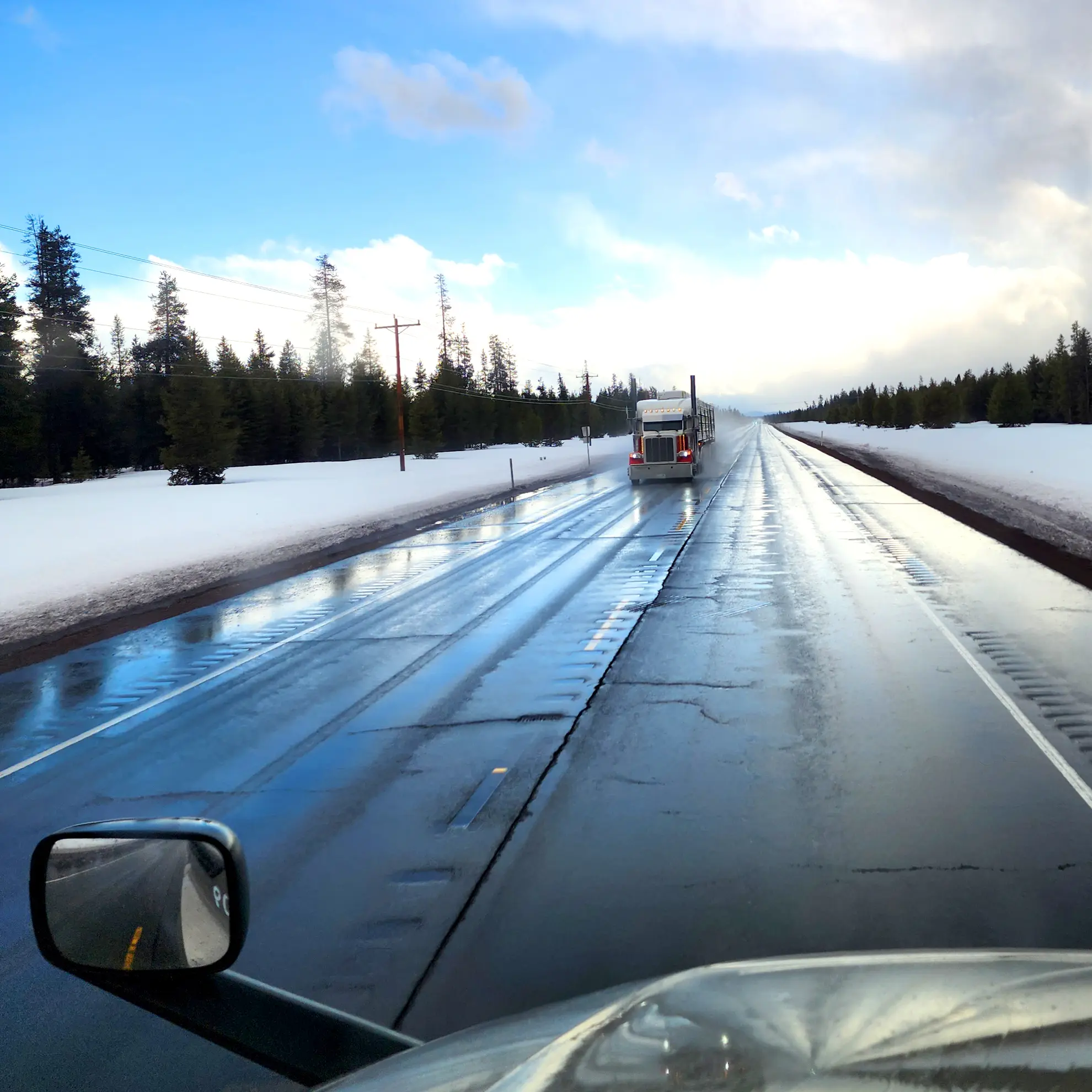 Un semirremolque equipado con un borrador automático circula por una carretera mojada rodeada de nieve y árboles bajo un cielo parcialmente nublado.