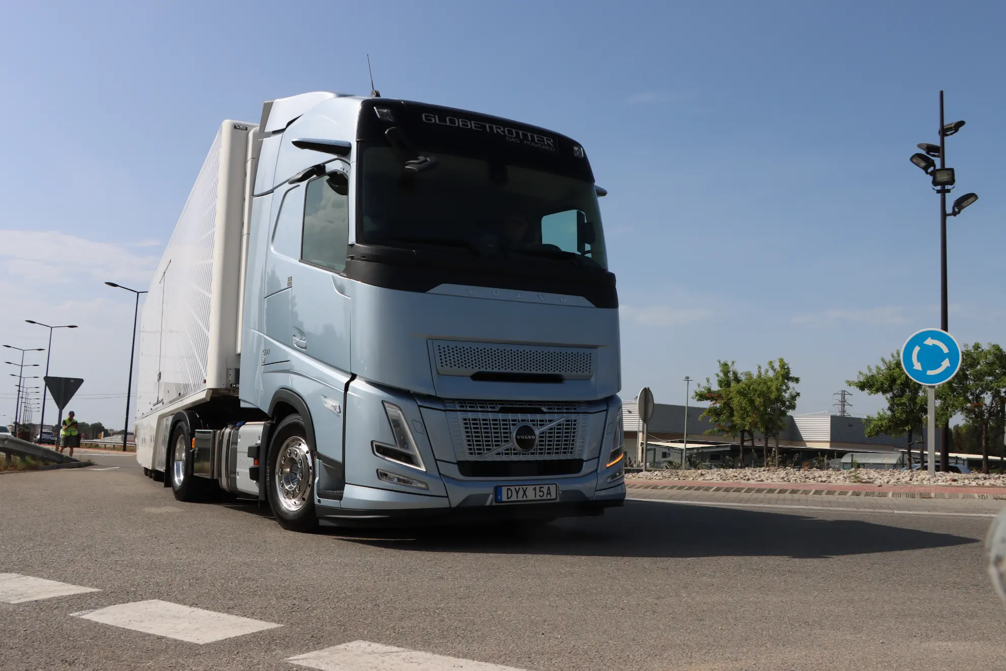 Un semirremolque Volvo a gas plateado circula por una carretera cercana a una rotonda bajo un cielo parcialmente nublado, ofreciendo a los gestores de flotas y a cualquier gestor de flotas una opción de transporte eficiente y ecológica.