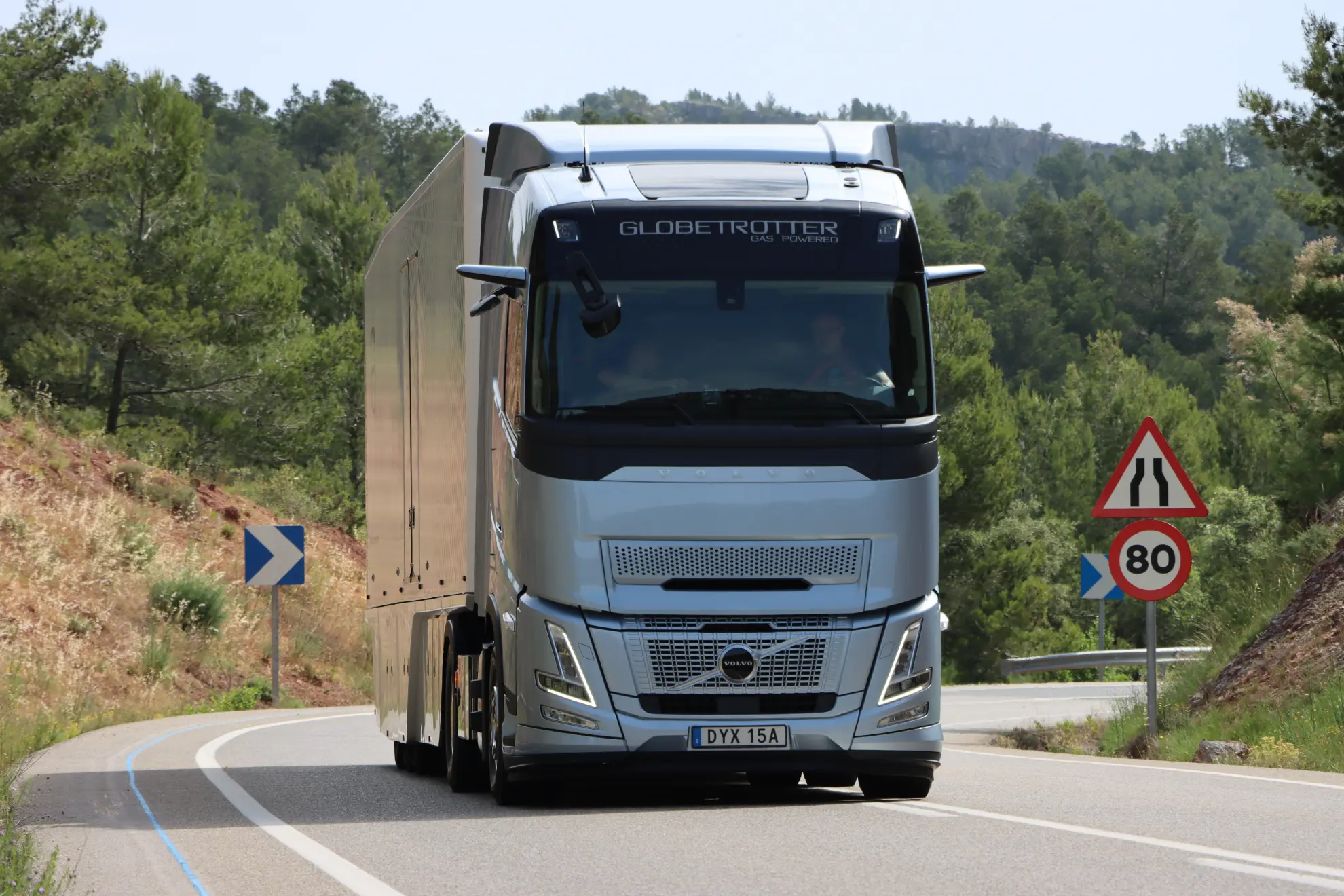 Un semirremolque Volvo plateado circula por una carretera sinuosa con señales de tráfico y árboles de fondo, mostrando la eficacia del Volvo a gas para quienes estén considerando soluciones de gas tco camión.