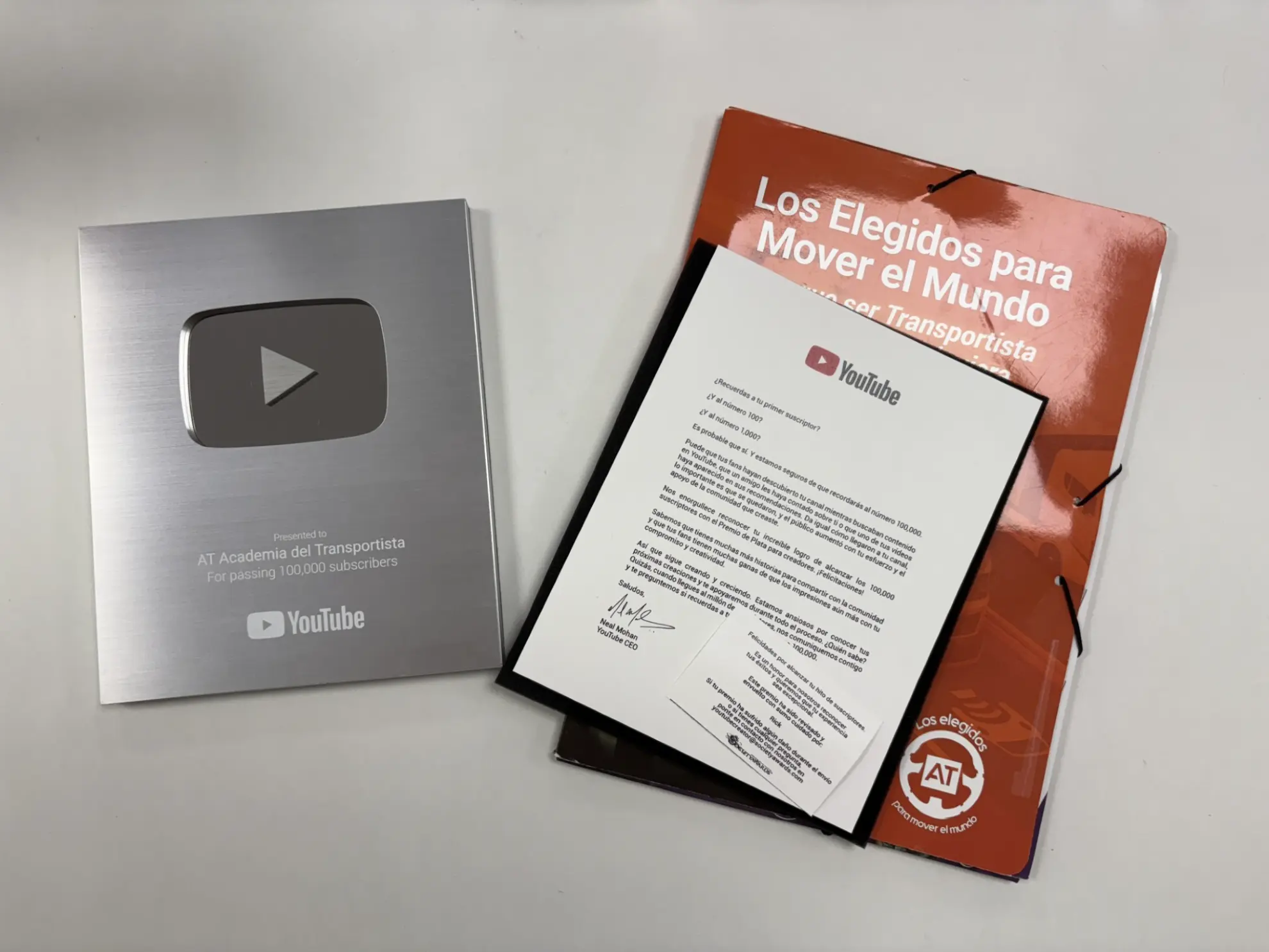 El premio YouTube Silver Play Button, una carta enmarcada, un folleto naranja y una hoja con la etiqueta "Borrador automático" descansan sobre una superficie blanca.