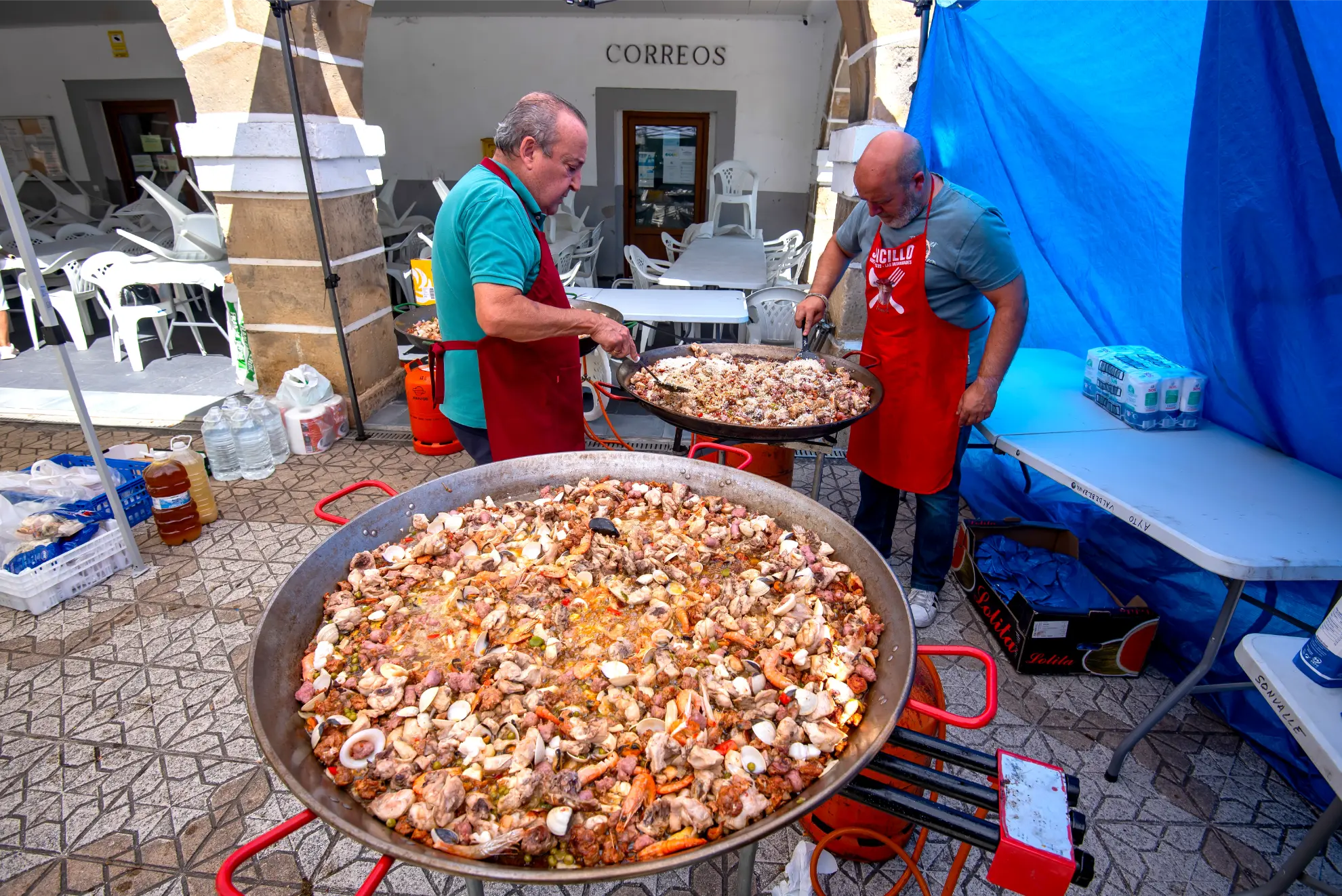 Dos hombres cocinan grandes sartenes de paella al aire libre bajo un toldo azul cerca de un edificio rotulado "Correos" durante la Quedada de Camiones 2025 en Camiones en Soncillo.