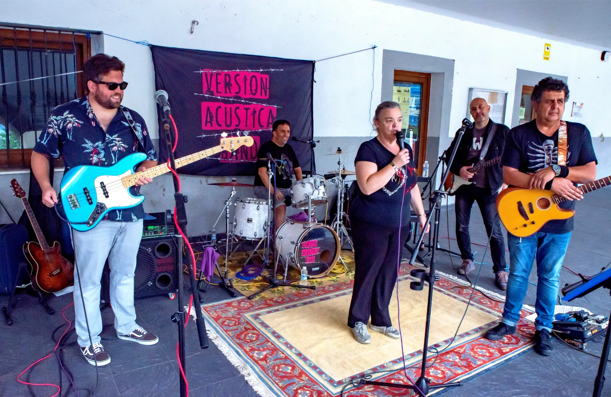 Cinco músicos con instrumentos tocan sobre una alfombra delante de una pancarta de "Version Acústica Band" en el interior de la Quedada de Camiones Soncillo 2025, perfecto para fotos memorables del animado evento.