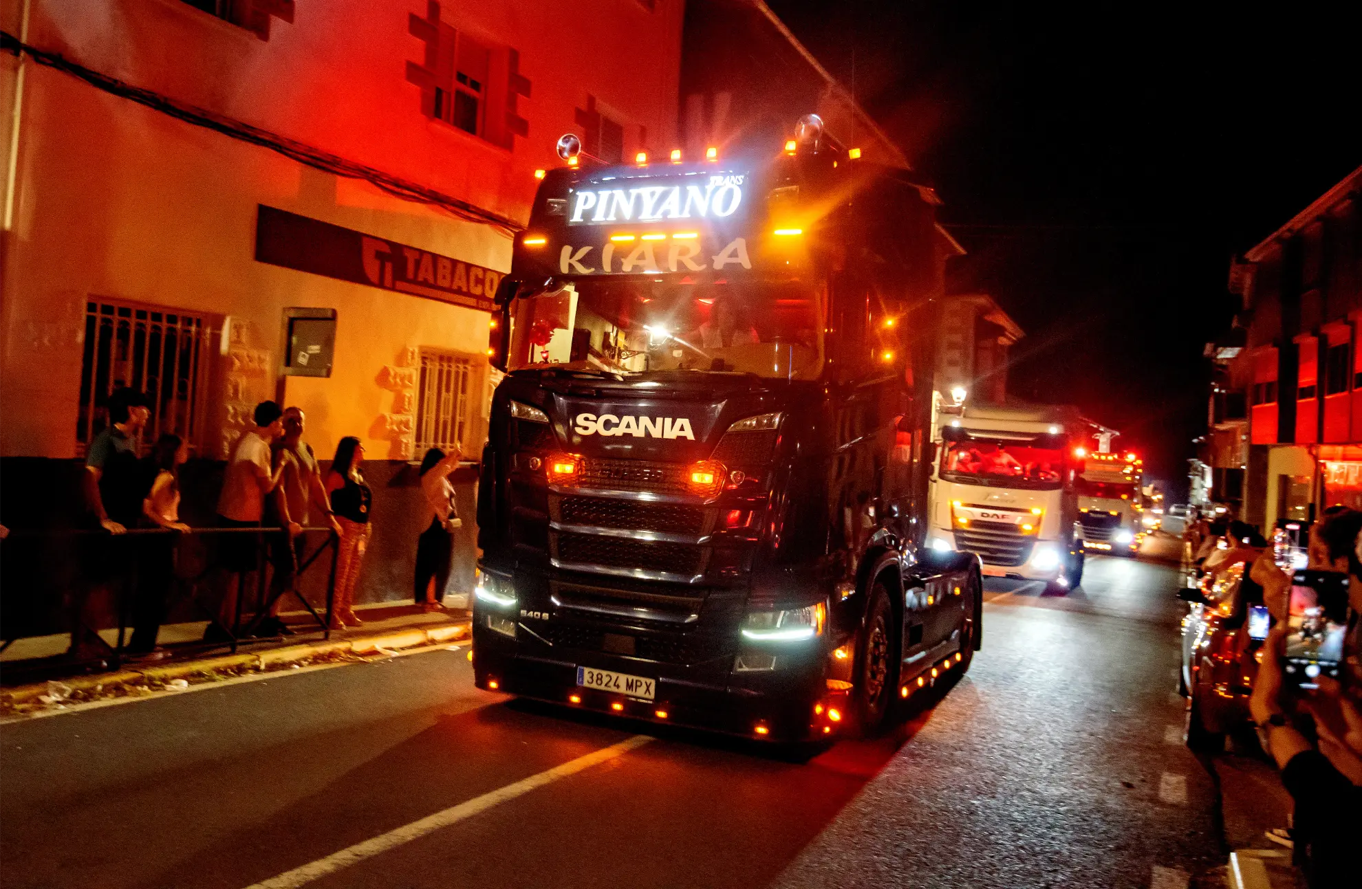 Un gran camión Scania iluminado recorre una calle por la noche durante el desfile de la Quedada de Camiones Soncillo 2025, observado por una multitud ansiosa por capturar fotos del deslumbrante espectáculo.