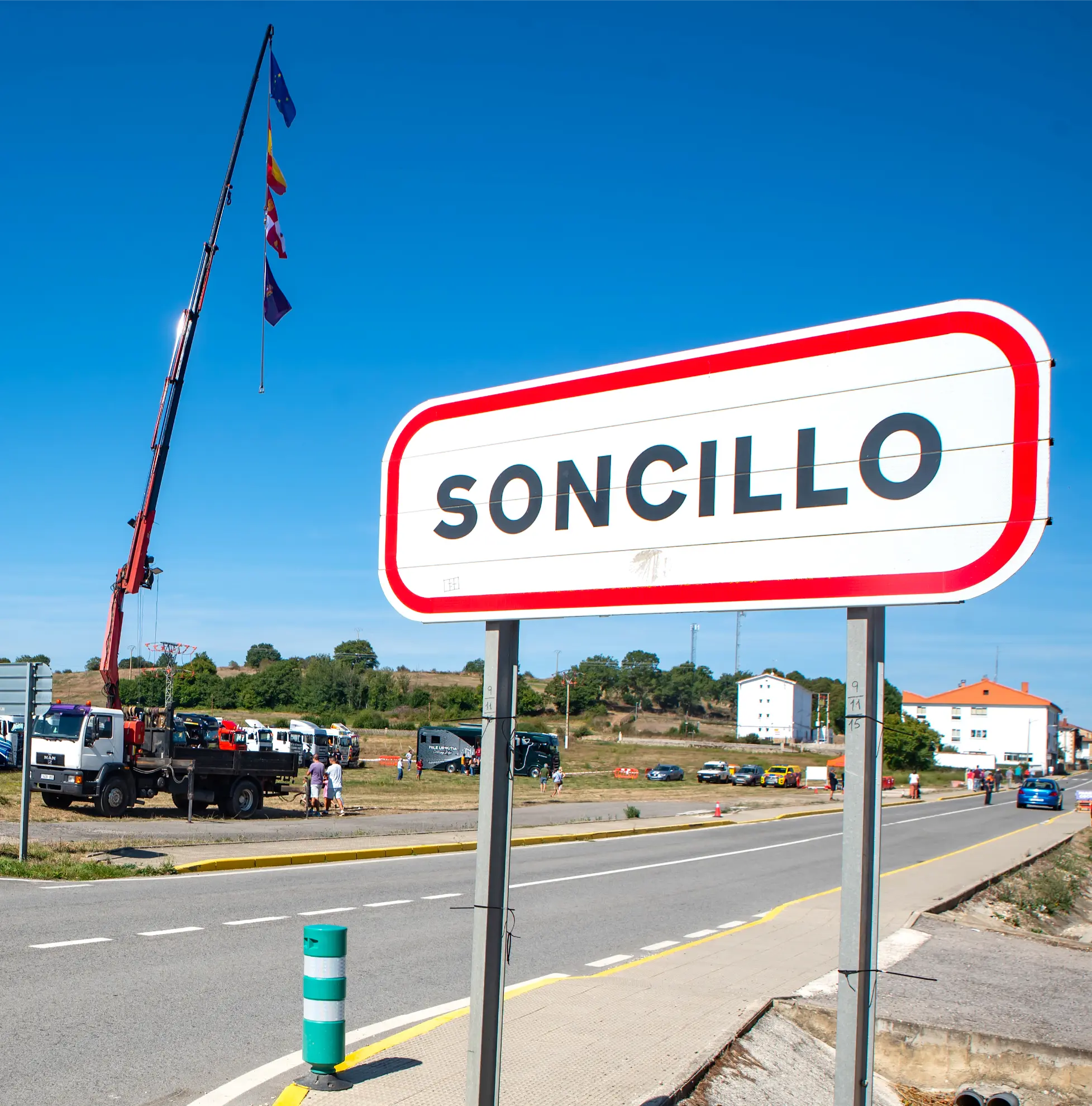 Una señal de tráfico de Soncillo con una grúa y banderas al fondo en un día soleado durante la Quedada de Camiones, parte de Crónica Soncillo 2025.