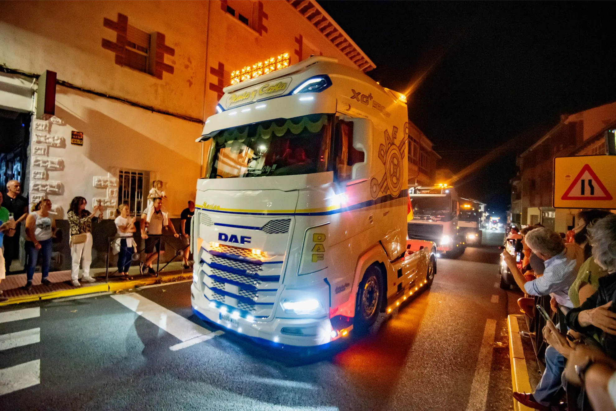 Un semirremolque DAF blanco con luces brillantes circula por una calle de noche durante la 2025 Quedada de Camiones en Soncillo, mientras la gente observa desde las aceras.