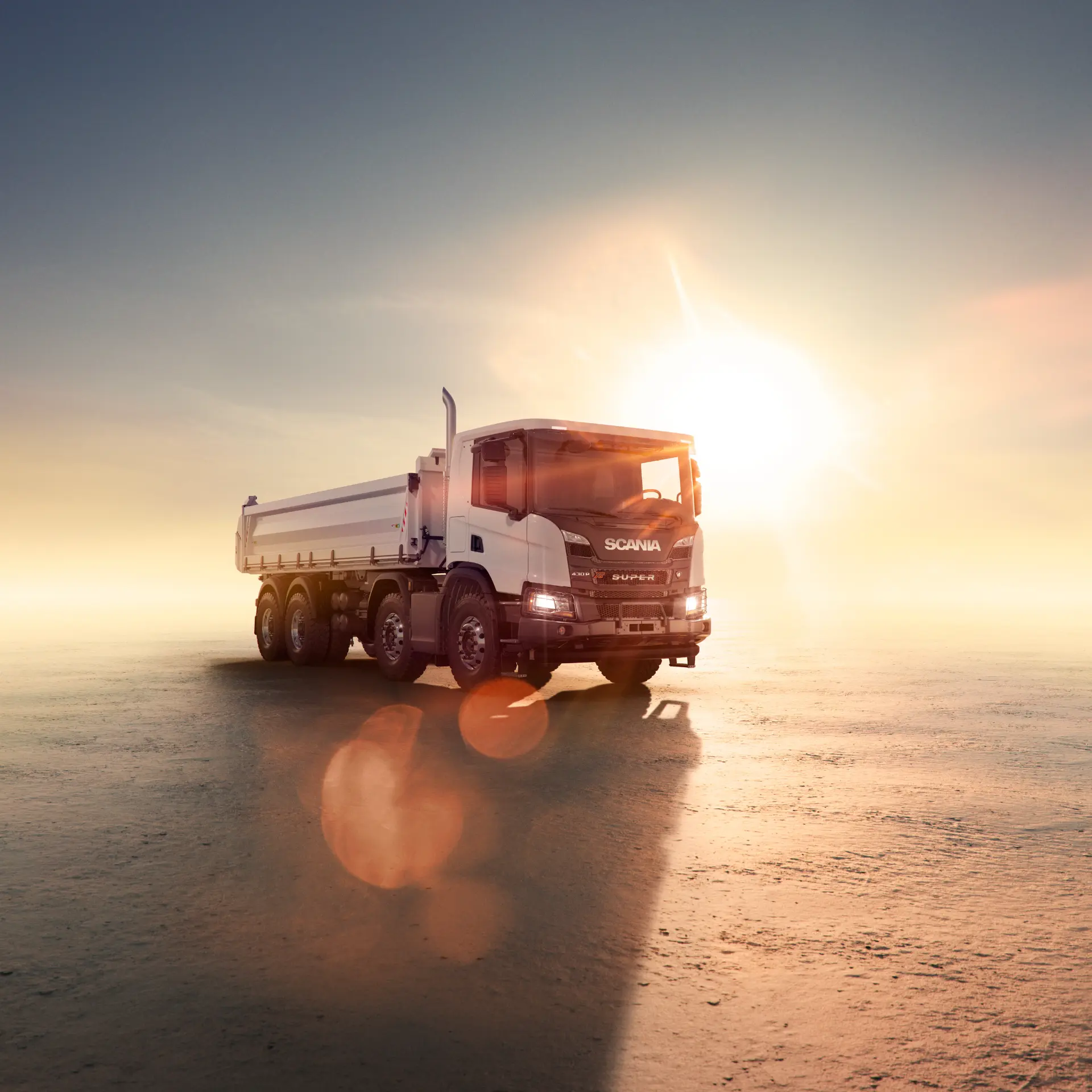 Un camión volquete Scania blanco, preparado para tareas de borrador automático, se encuentra sobre una superficie plana y vacía con el sol bajo en el horizonte.
