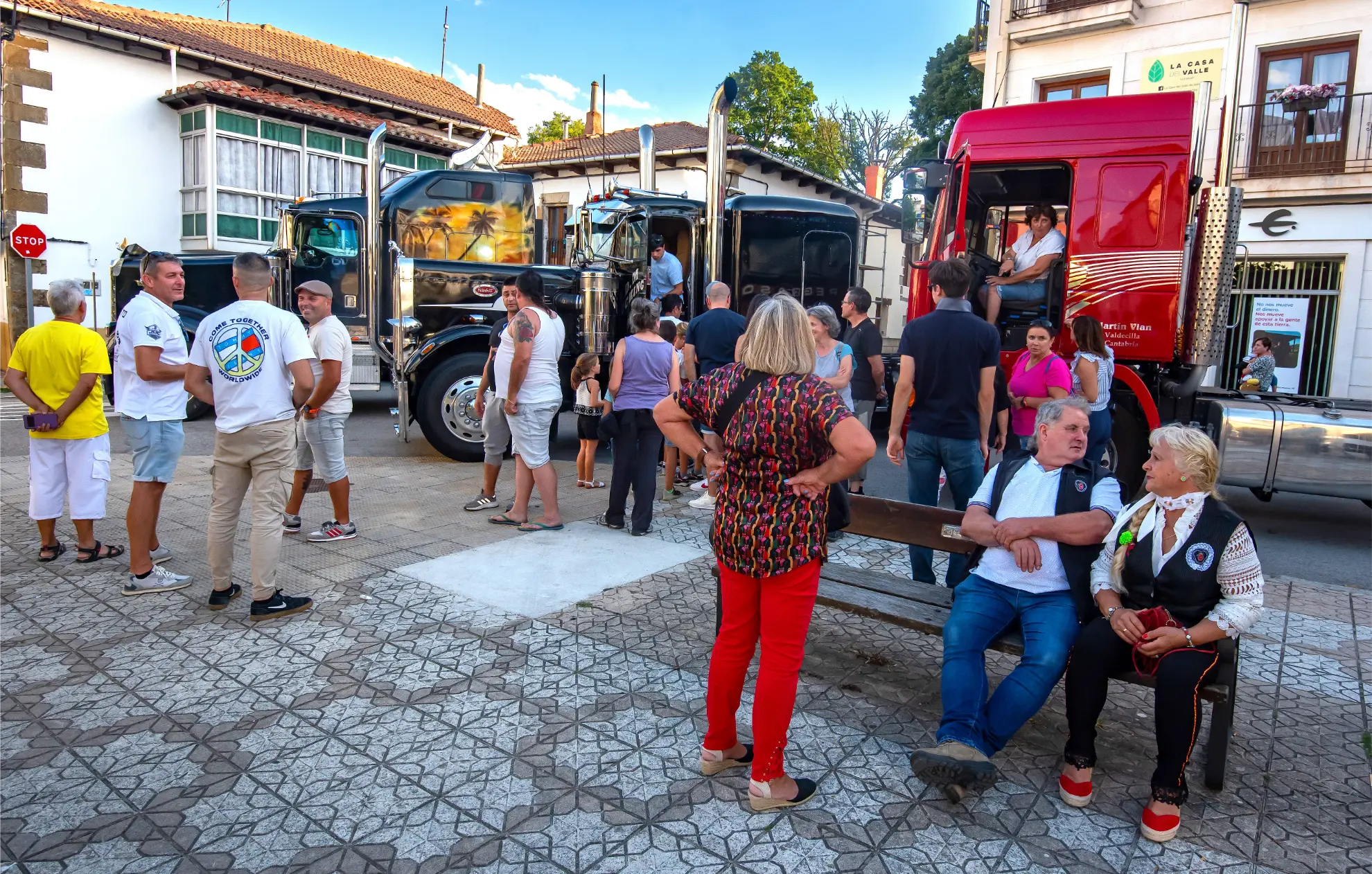 La gente se reúne cerca de grandes camiones en una animada plaza del pueblo en un día soleado para la Quedada de Camiones en Soncillo 2025, algunos sentados y otros de pie mientras disfrutan del vibrante ambiente.