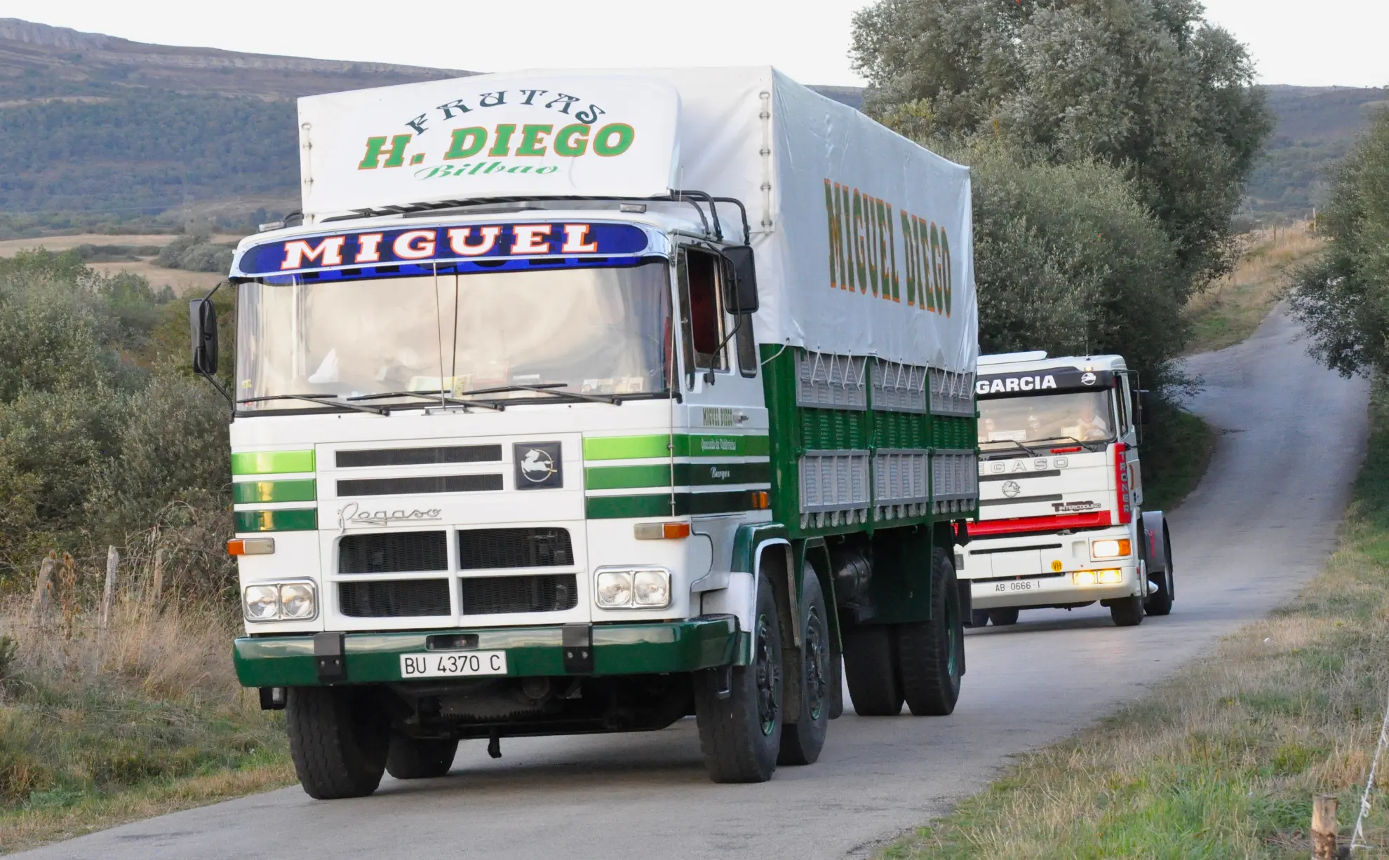 Dos camiones antiguos con letreros de colores circulan por un estrecho camino rural rodeado de vegetación durante la 2025 Quedada de Camiones en Soncillo.