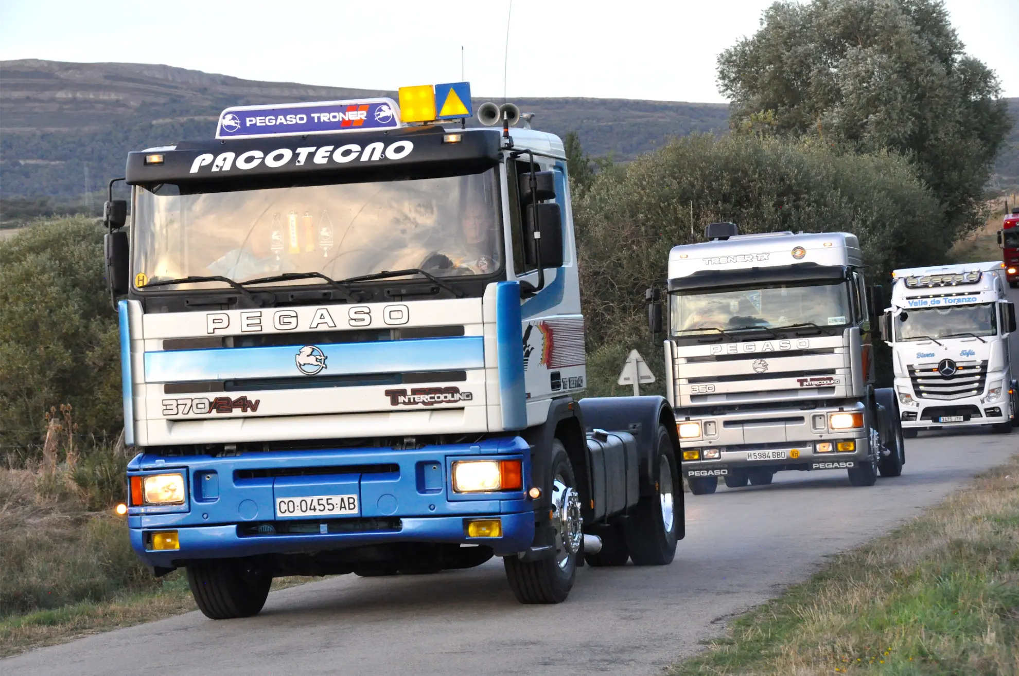 Tres camiones Pegaso azules y blancos circulan por un camino rural con árboles y colinas de fondo, captando un momento de la crónica de la 2025 Quedada de Camiones Soncillo.