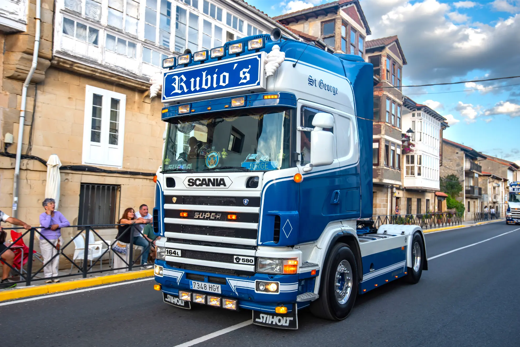 Un camión Scania azul y blanco rotulado "Rubio's" circula por una calle llena de gente y casas, en la colección de fotos Camiones en Soncillo 2025.