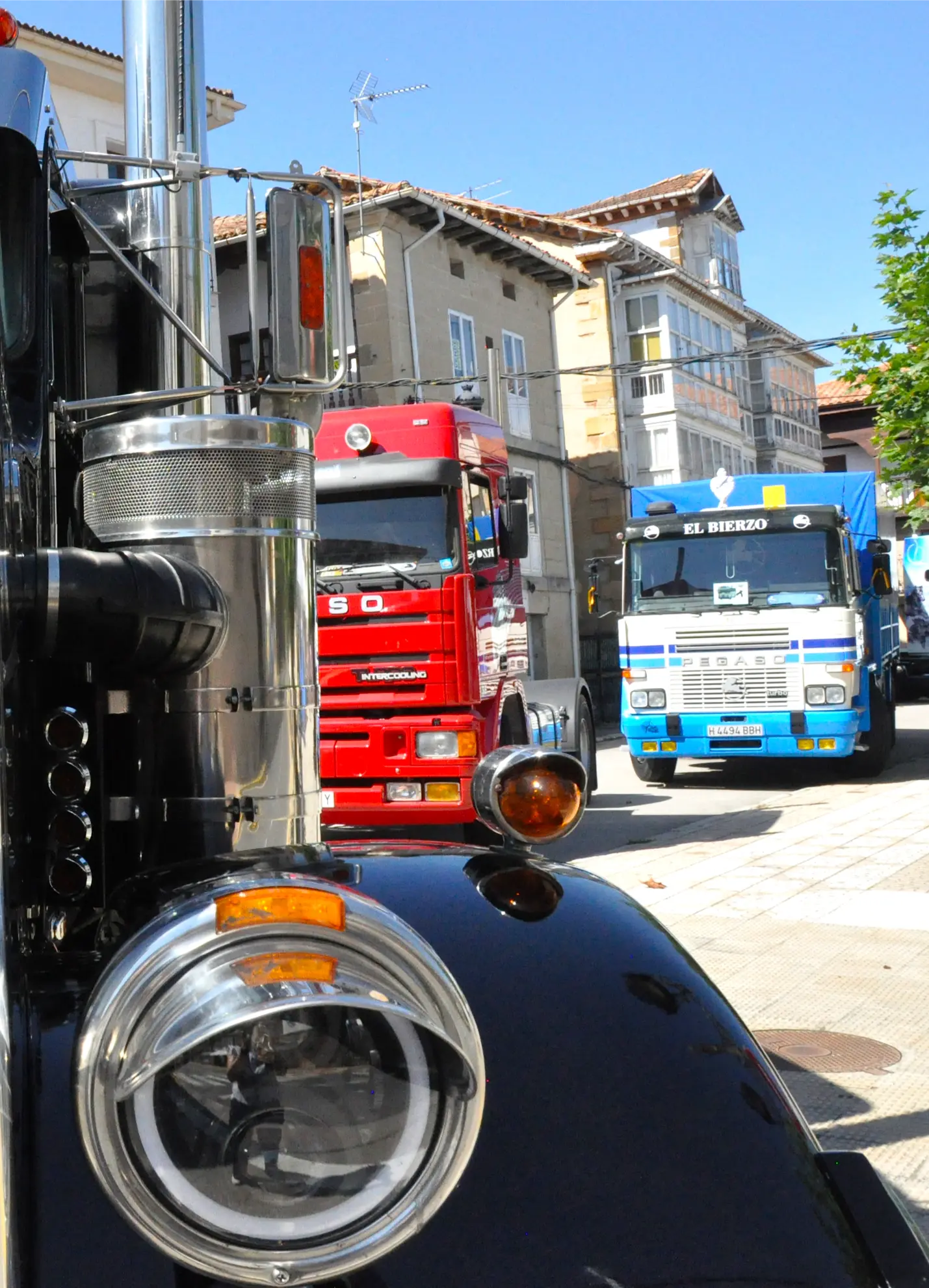 Primer plano de un faro de camión negro con camiones rojos y azules aparcados en una calle soleada durante la 2025 Quedada de Camiones en Soncillo.