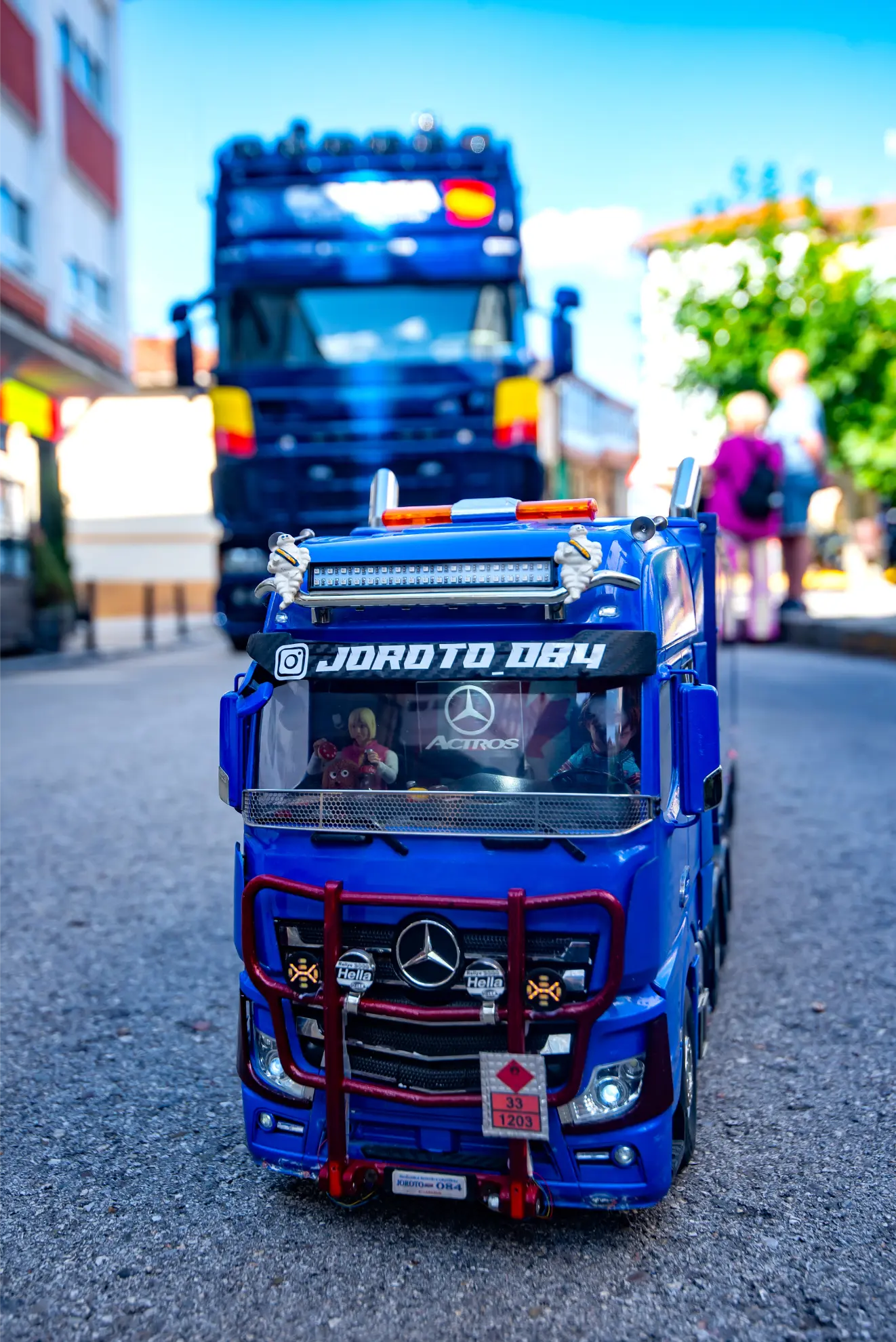 Un camión Mercedes azul de juguete se sienta en la calle, haciendo eco de los camiones reales de la Quedada de Camiones Soncillo 2025, con un camión azul más grande y edificios al fondo, captando un momento digno de una crónica.