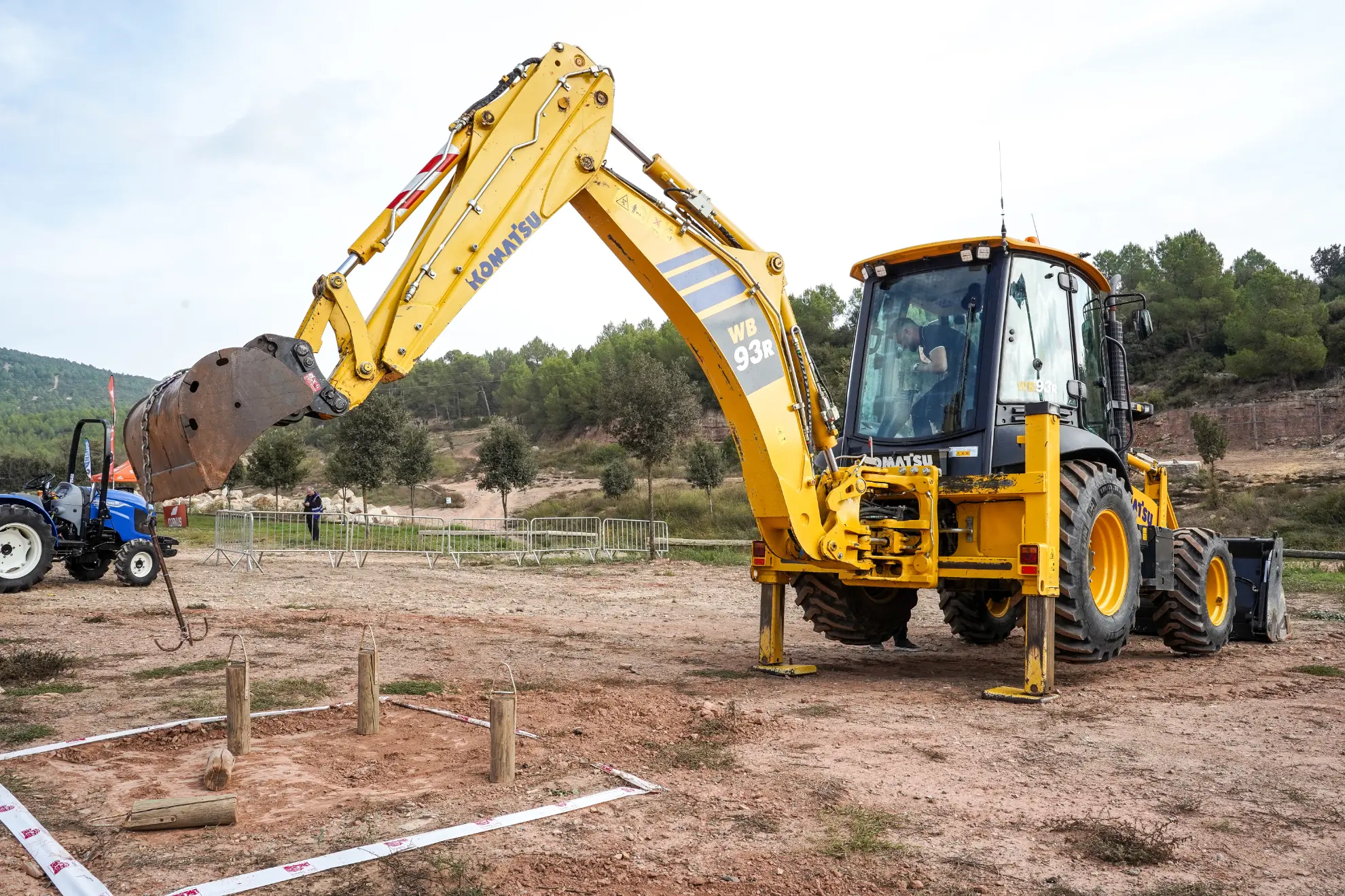 Una excavadora amarilla mueve troncos en un campo de tierra, rodeado de árboles y vallas en el concesionario Renault Trucks Lleida.