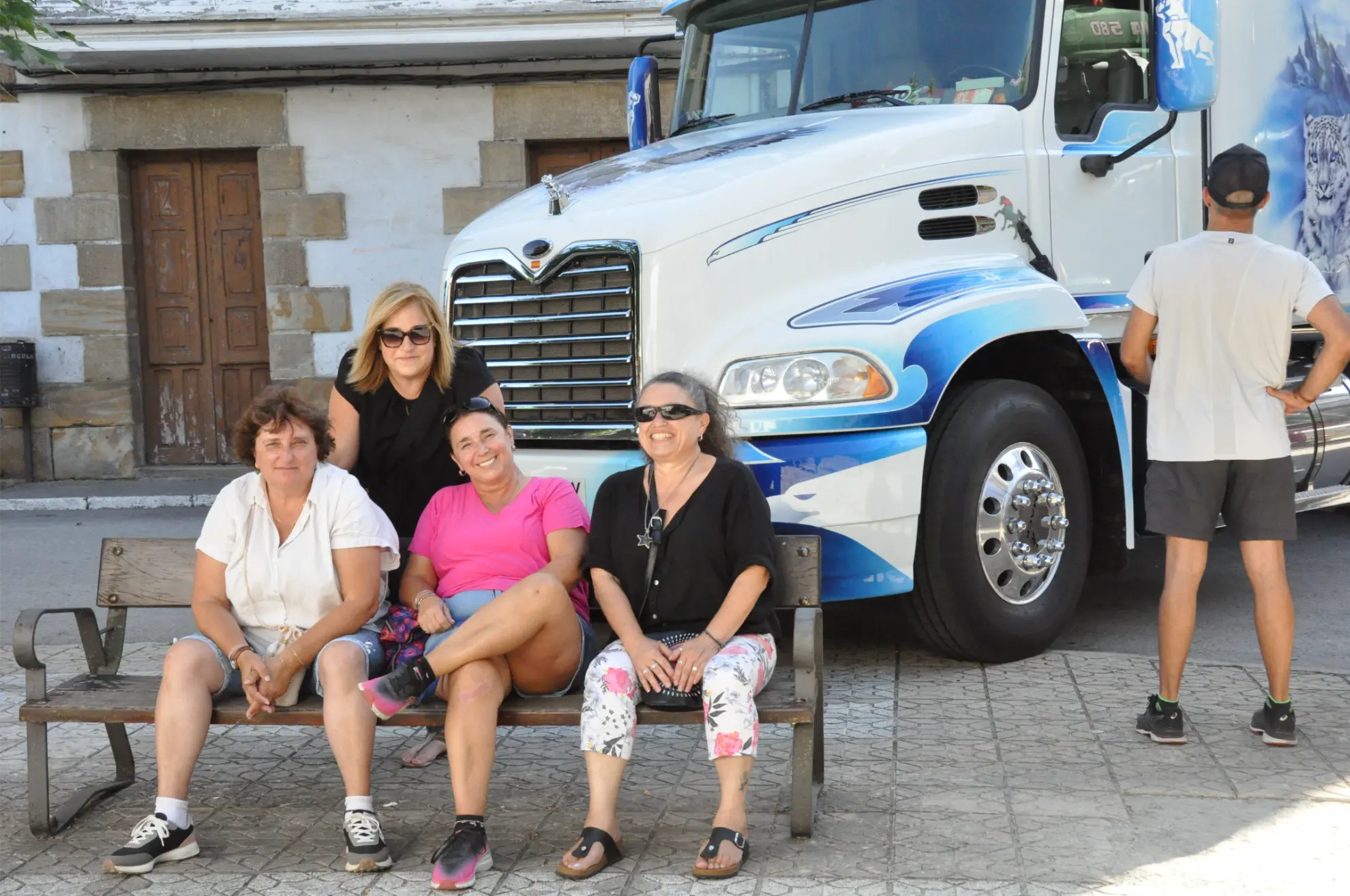 Cuatro mujeres se sientan sonrientes en un banco frente a un gran camión blanco y azul en Camiones en Soncillo; un hombre permanece cerca, captando un momento alegre de esta crónica de la Quedada 2025.