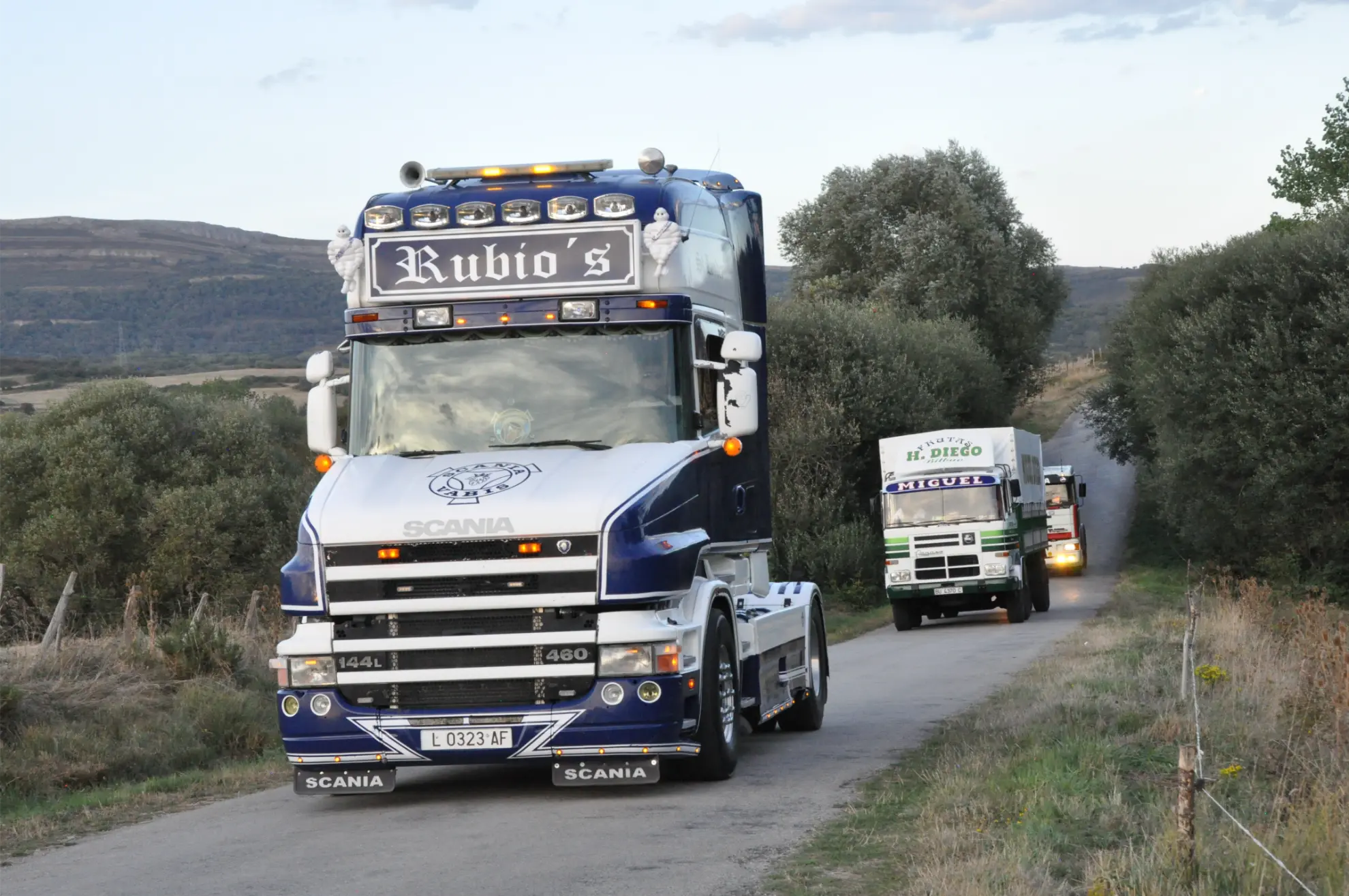 Tres coloridos semirremolques circulan por una estrecha carretera rural rodeada de hierba, árboles y colinas lejanas durante el Soncillo 2025 Quedada de Camiones.