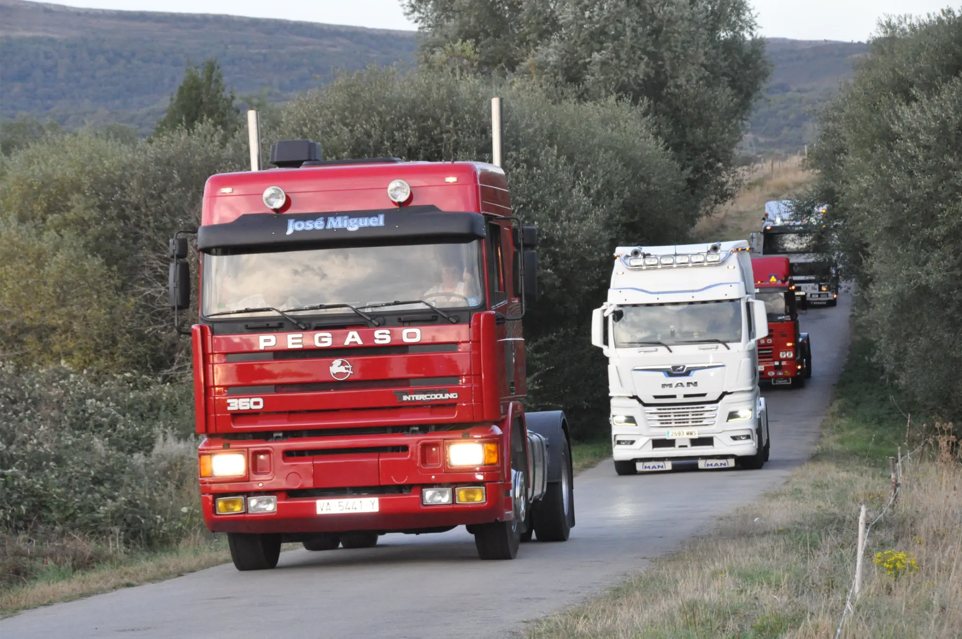 Un camión Pegaso rojo encabeza un convoy durante la Quedada de Camiones en una estrecha carretera rural bordeada de árboles: una de las muchas fotos memorables de Soncillo 2025.
