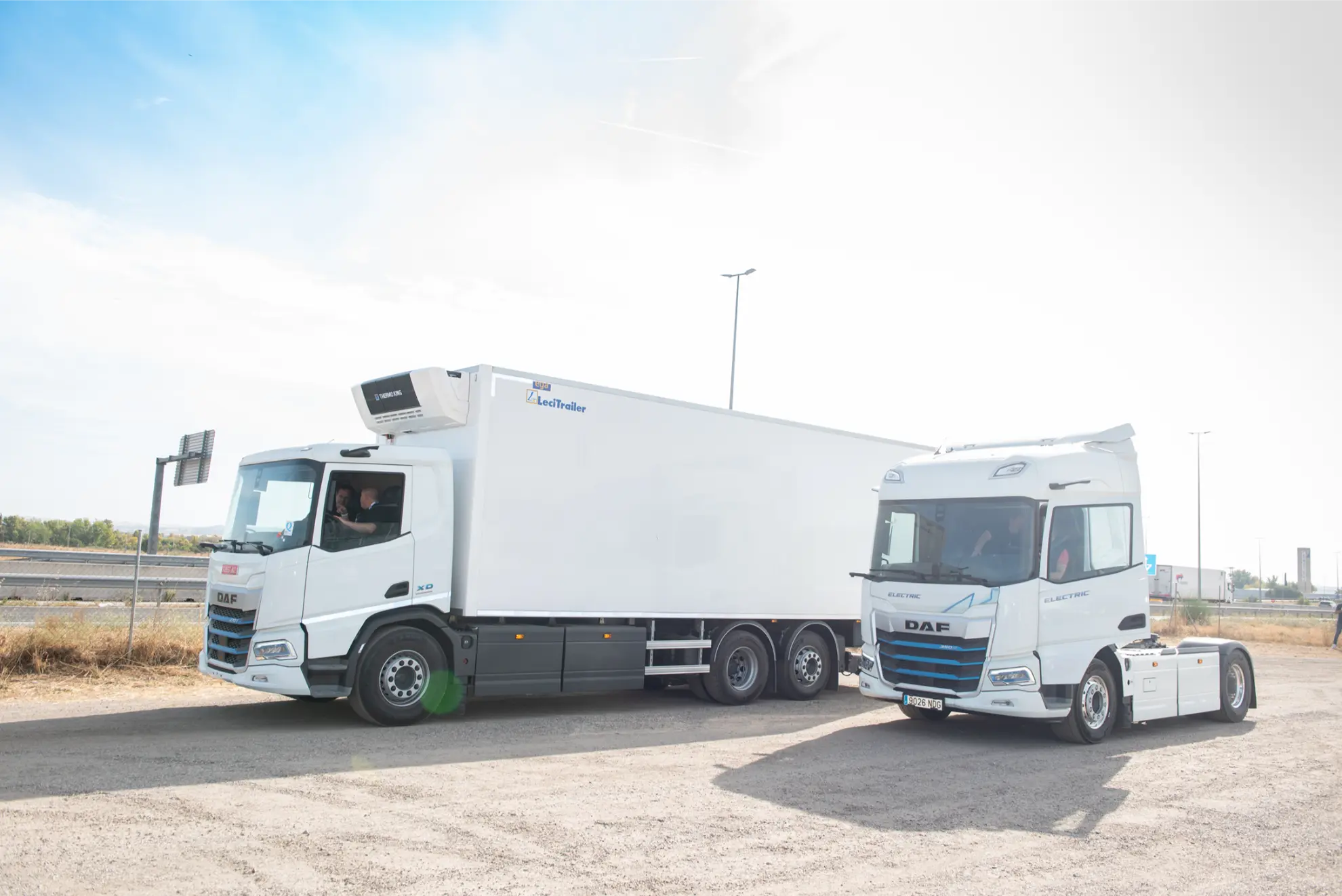 Dos camiones DAF blancos aparcados en un camino de tierra bajo un cielo brillante y parcialmente nublado, equipados con la última tecnología de borrador automático.