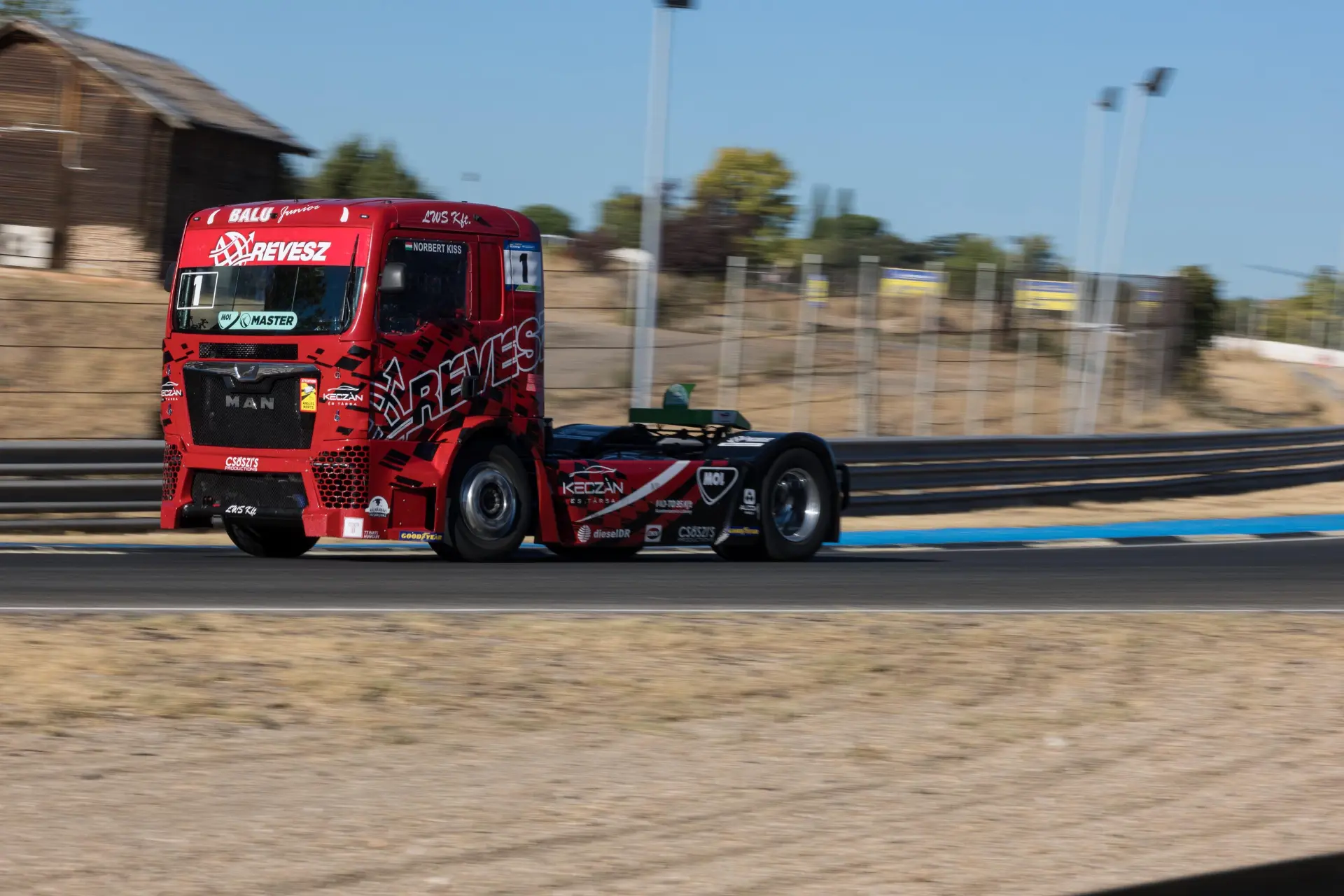 Un camión de carreras rojo con borrador automático acelera en una curva de una pista de carreras bajo un cielo azul despejado.