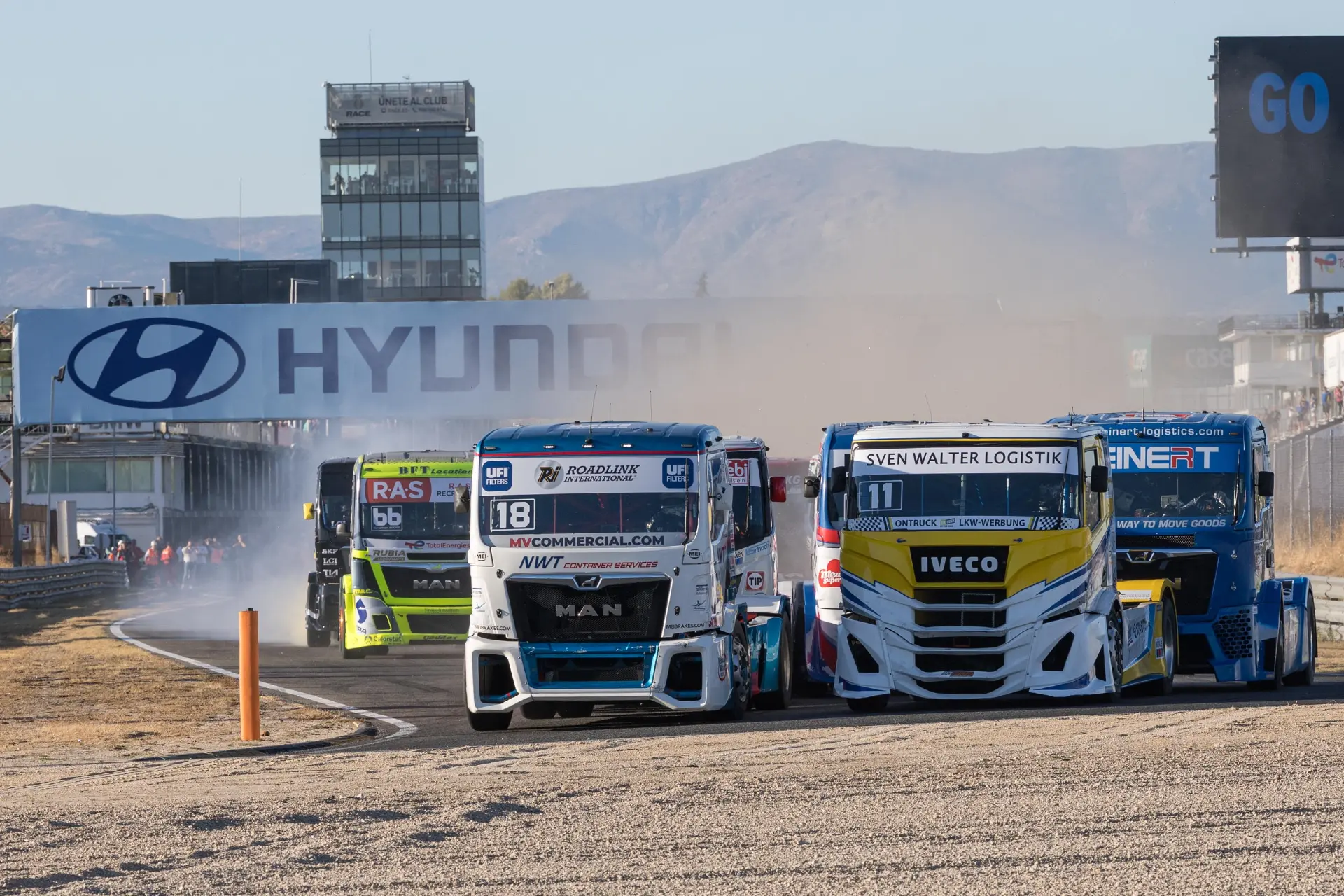 Cuatro camiones de carreras toman una curva en una pista de carreras, levantando polvo con la señalización de Hyundai de fondo, mientras el borrador automático capta cada momento emocionante.