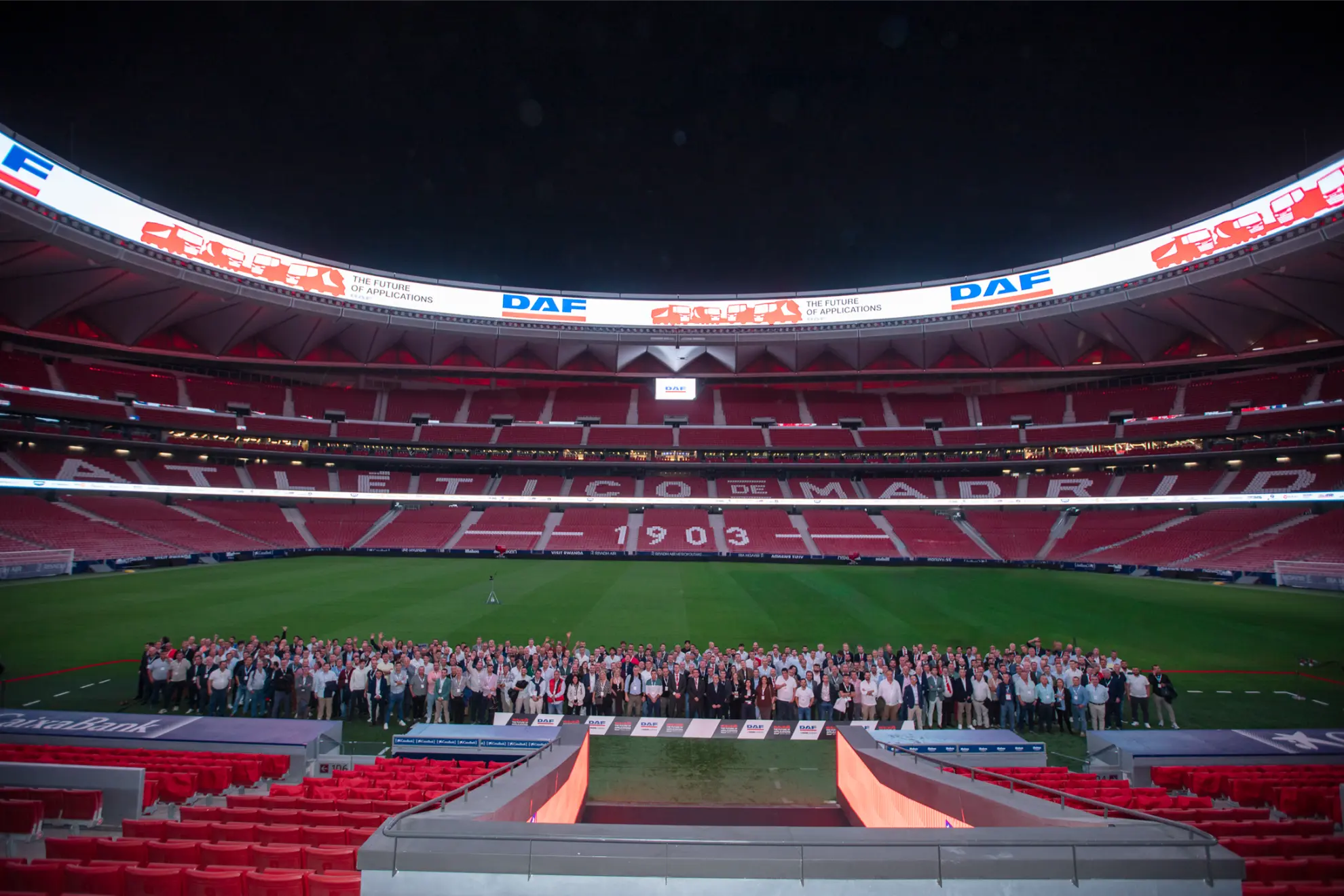 Un numeroso grupo de personas posa para una foto en el campo de un estadio del Atlético de Madrid vacío por la noche.