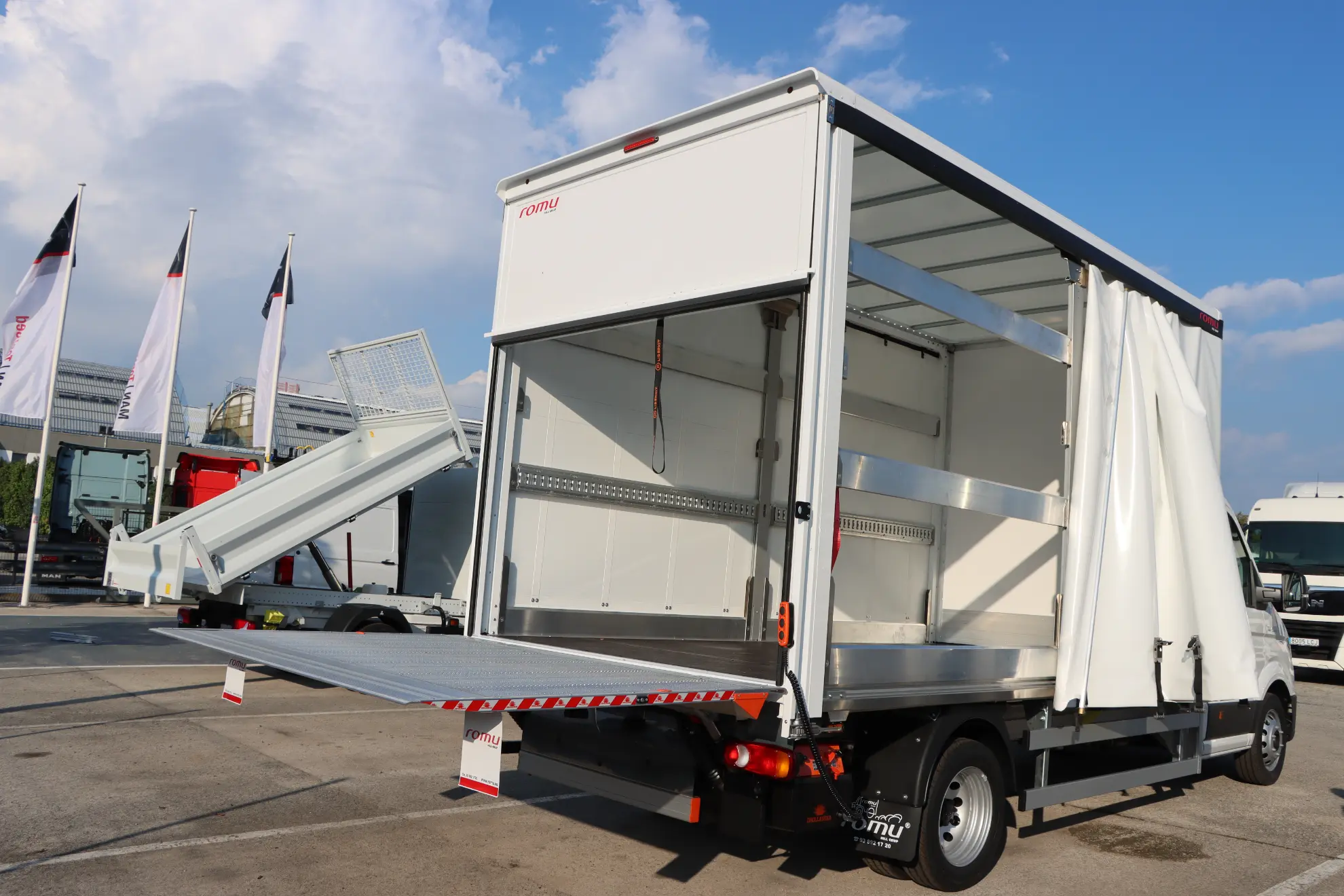 Un camión MAN TGE blanco con caja, ideal entre los vehículos para autónomos, con la puerta trasera y la cortina lateral abiertas, está aparcado en un solar al aire libre.