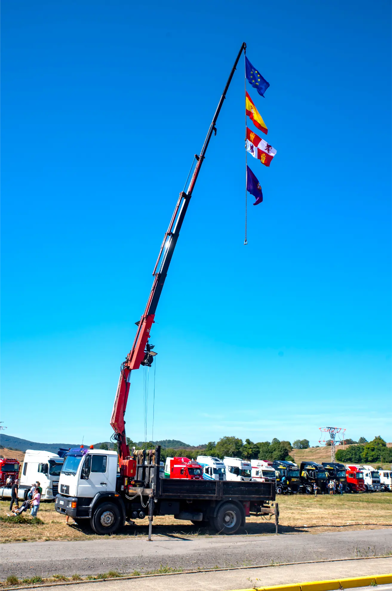 Un camión grúa iza cuatro banderas de colores por encima de un campo cubierto de hierba en la Quedada de Camiones 2025, con coches y gente reunidos para Camiones en Soncillo.
