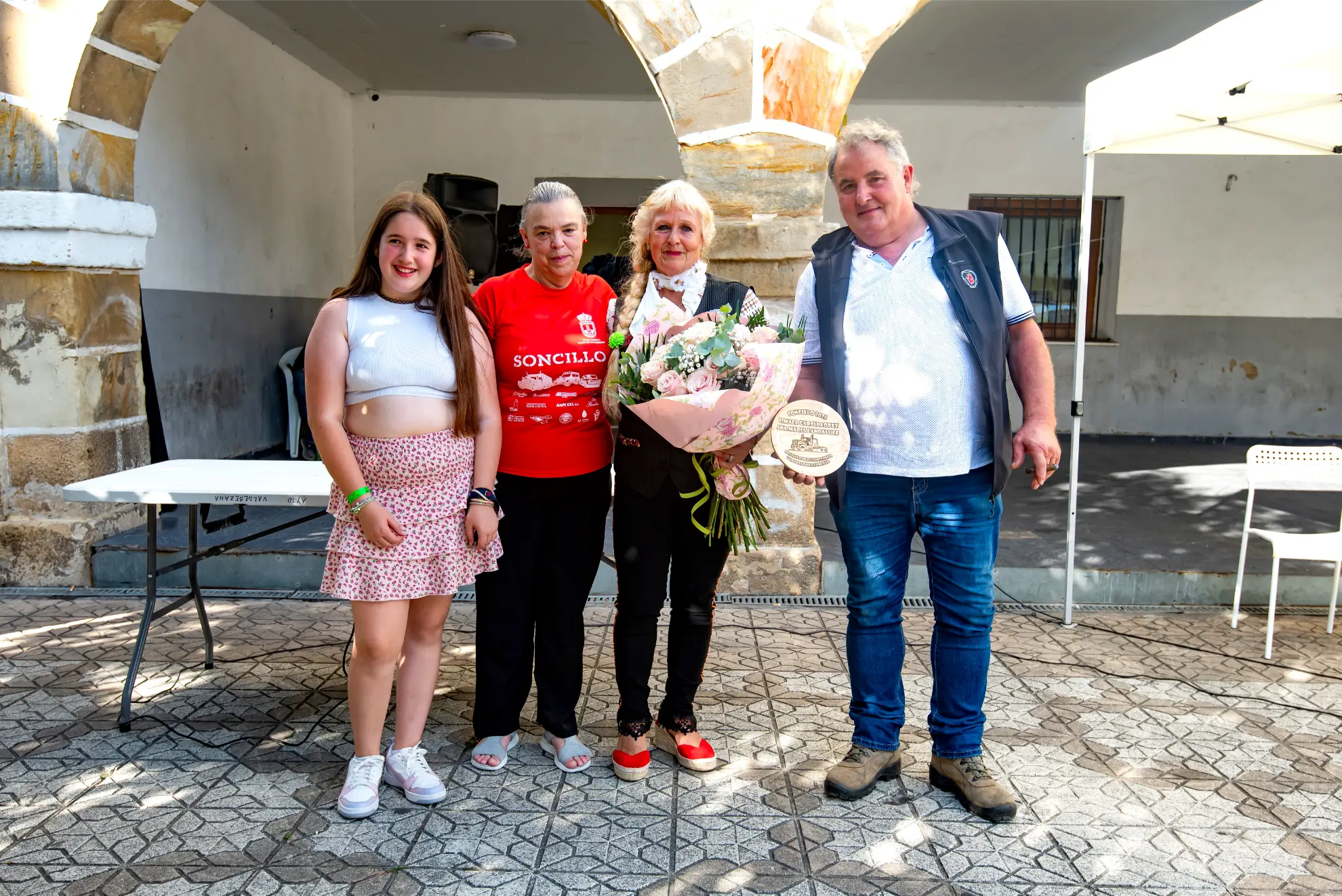 Cuatro personas permanecen juntas al aire libre en la Quedada de Camiones Soncillo 2025, una de ellas con un ramo de flores y una placa, todas sonriendo para las fotos.
