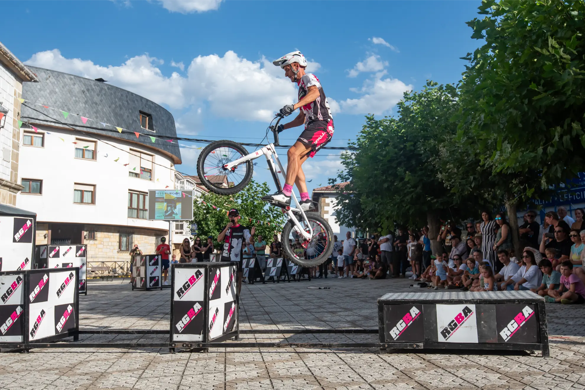 Un ciclista realiza un truco, saltando una bicicleta por encima de obstáculos ante la multitud durante la 2025 Quedada de Camiones en la plaza del pueblo de Soncillo.
