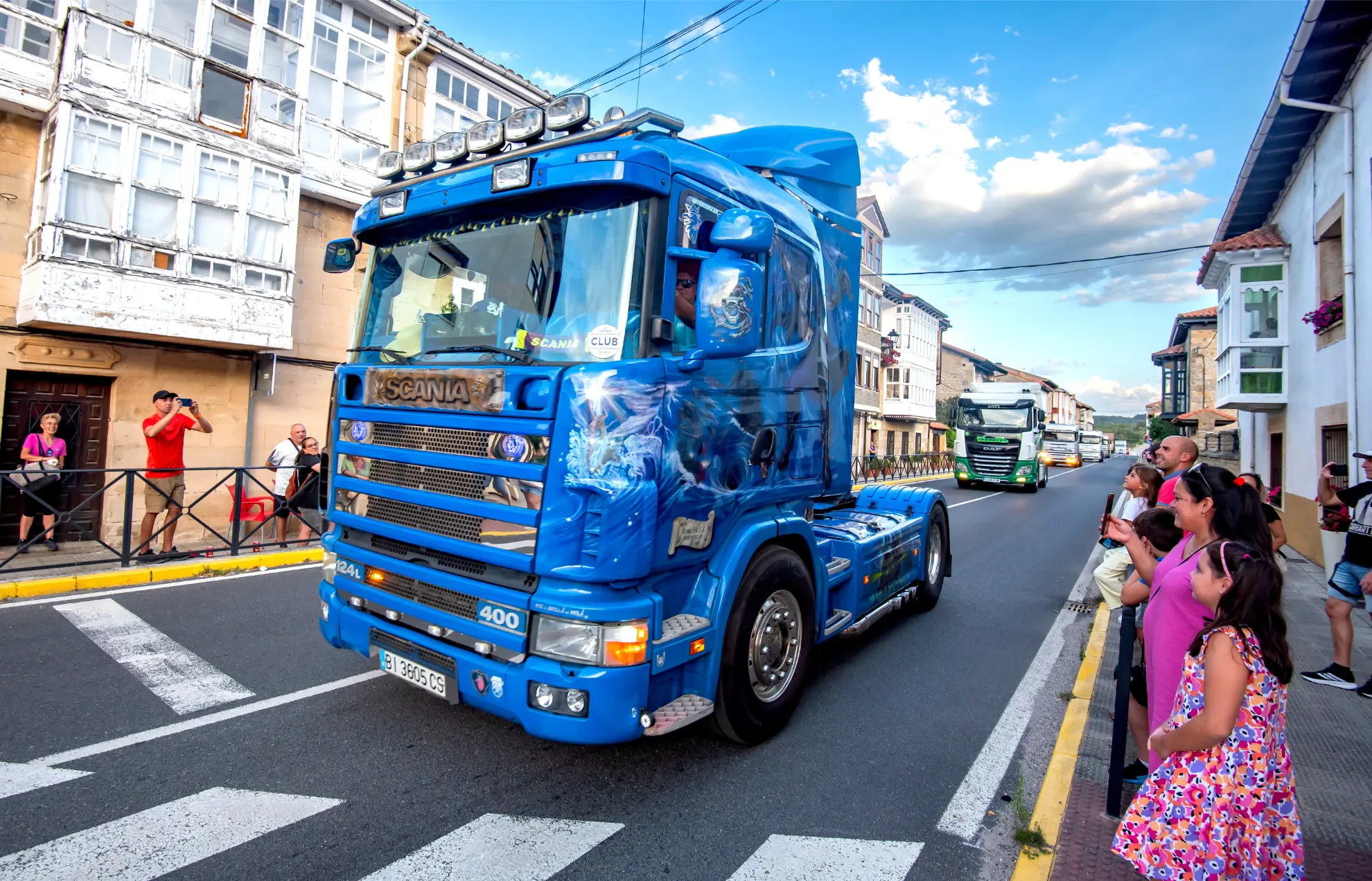 Un camión Scania azul brillante atraviesa la ciudad durante la Quedada de Camiones 2025, mientras la gente en la acera hace fotos y observa.
