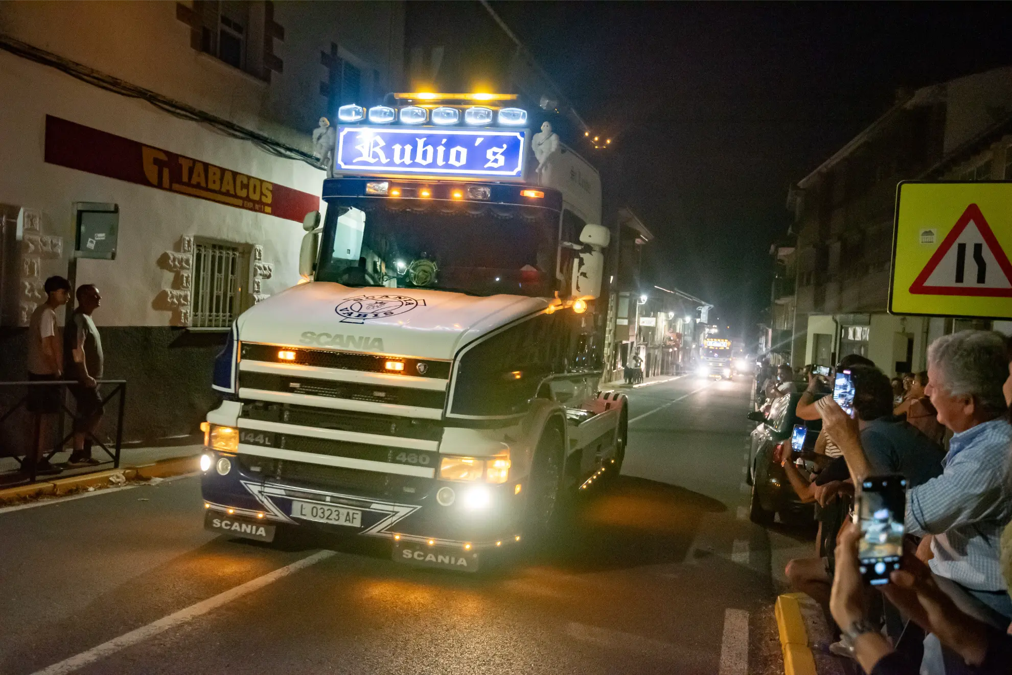 Un gran camión con luces brillantes circula por una calle de noche durante la Quedada de Camiones 2025 en Soncillo, observado por la gente que hace fotos.
