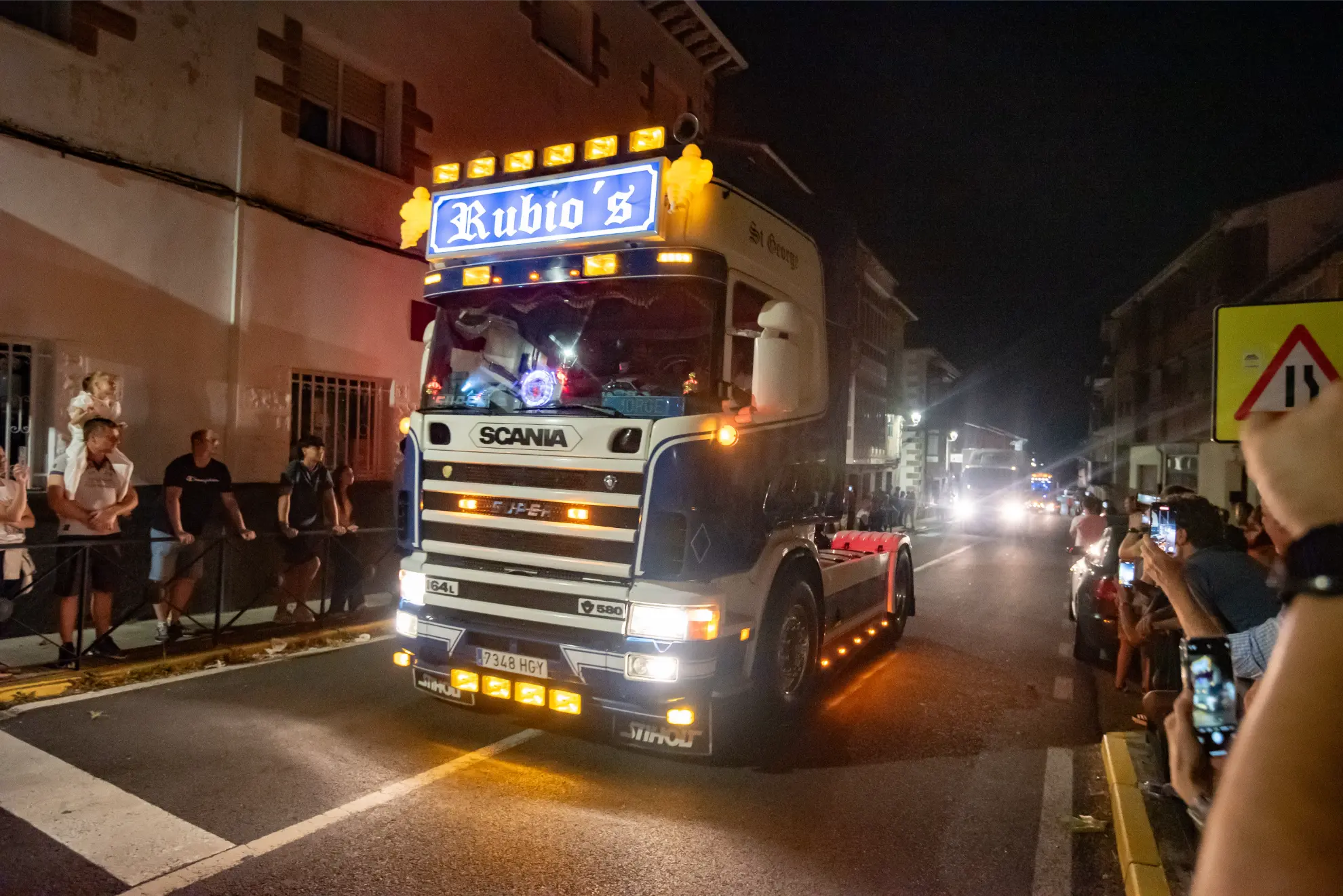 Un camión decorado con el rótulo "Rubio's" atraviesa una calle por la noche durante la Quedada de Camiones Soncillo 2025, observado por gente de ambos lados: una auténtica crónica de celebración festiva.