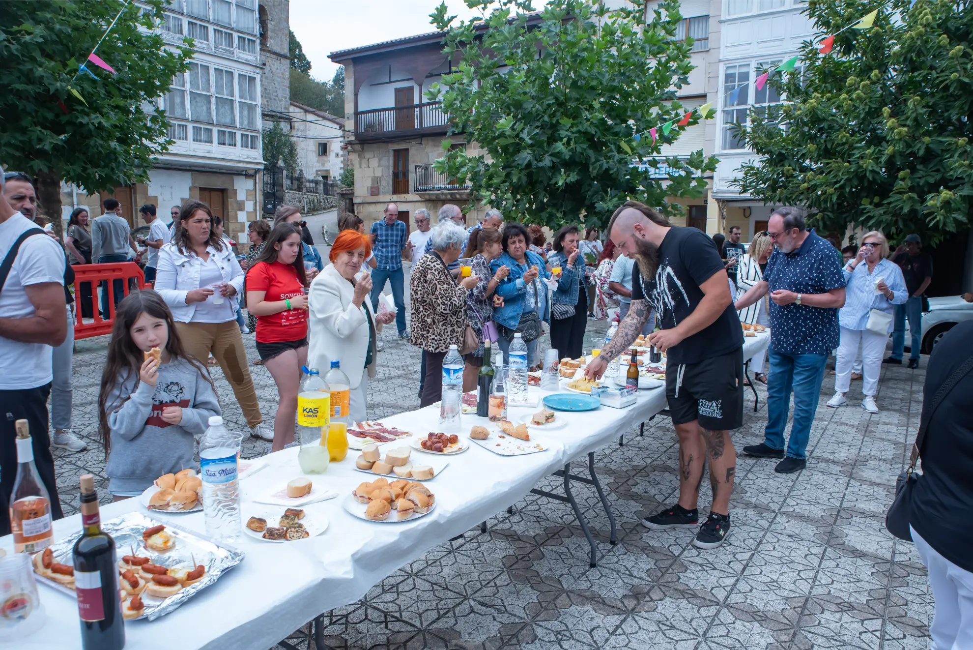 La gente se reúne al aire libre alrededor de mesas con comida y bebida, disfrutando de un acto comunitario en una plaza embaldosada durante la 2025 Quedada de camiones en Soncillo.
