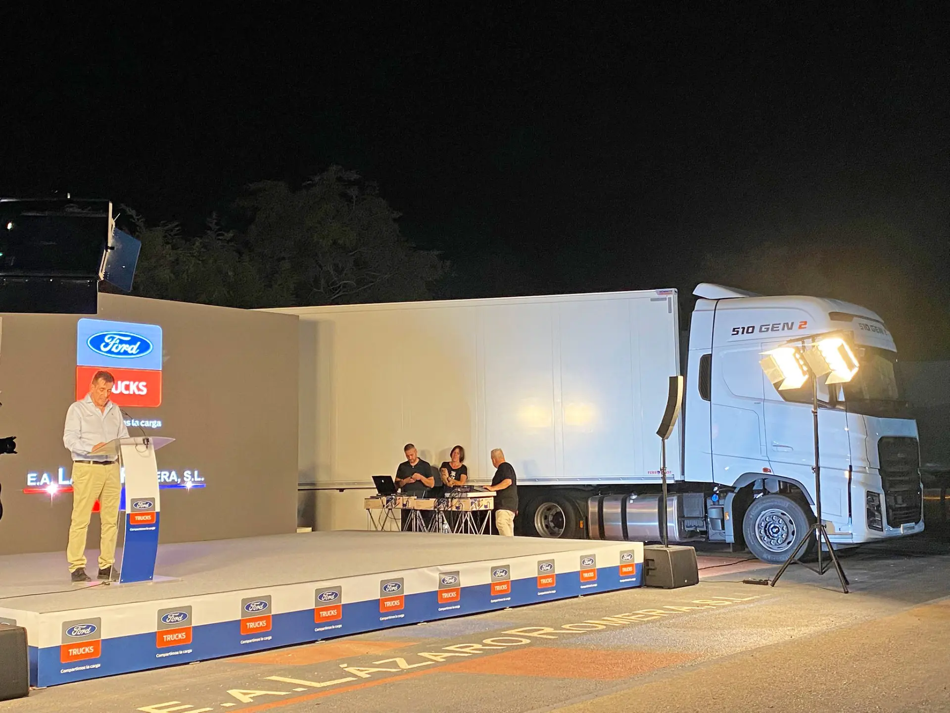 Un hombre habla en el escenario de un evento de Ford Trucks al aire libre por la noche en Murcia; la gente se sienta en una mesa junto a un gran camión blanco F-MAX GEN2.