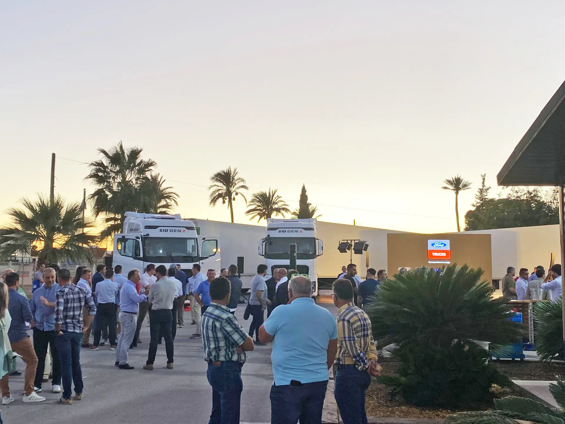 Un grupo de personas se reúne al aire libre cerca de dos grandes Ford Trucks al atardecer en Murcia, con las palmeras silueteadas al fondo.