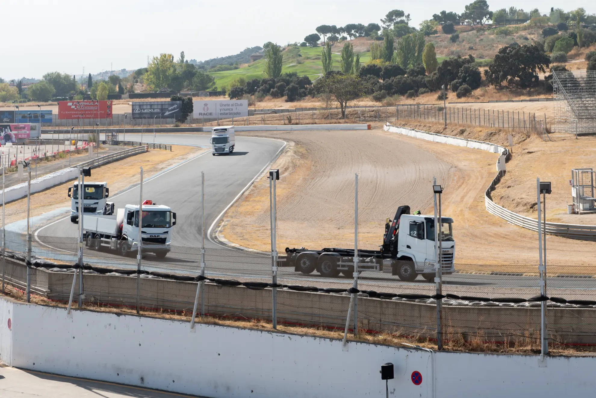Tres camiones blancos circulan por una pista asfaltada rodeada de hierba seca y gradas vacías.