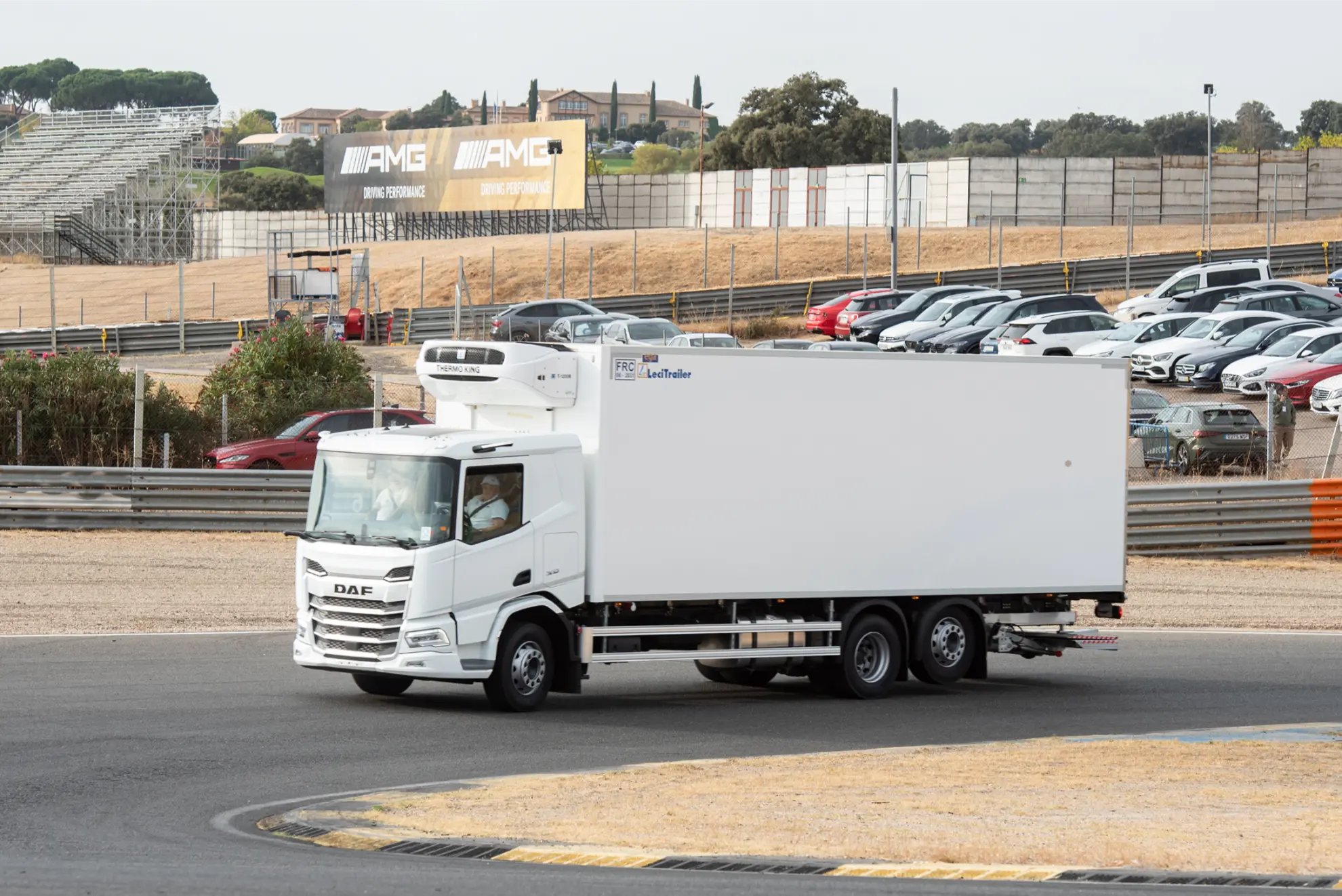 Un camión DAF refrigerado blanco, equipado con un borrador automático, circula por un circuito de carreras cerca de coches aparcados y tribunas.