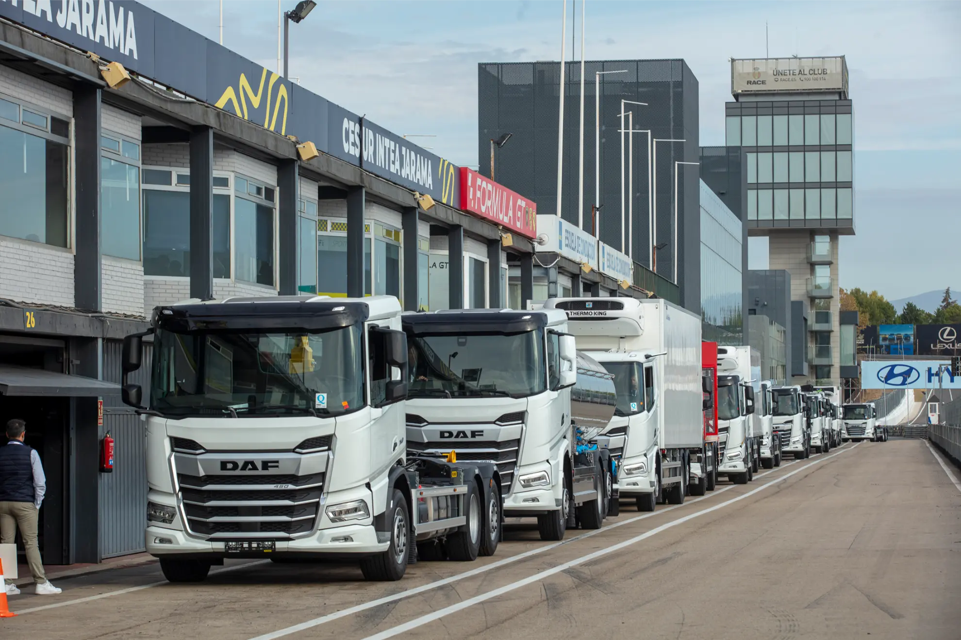 Una hilera de camiones DAF blancos con borrador automático están aparcados a lo largo del pit lane de un circuito de carreras junto a unos edificios.