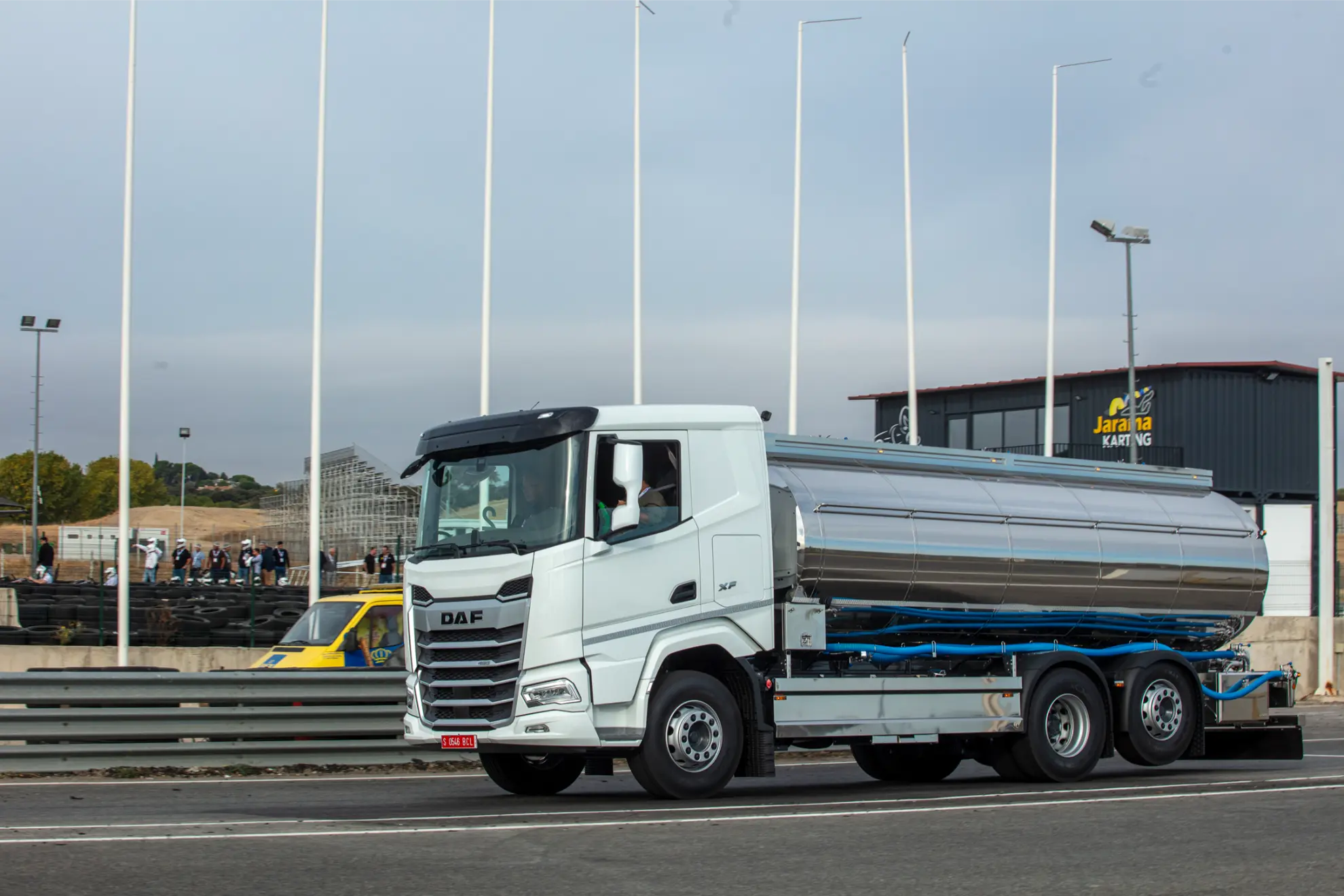 Un camión cisterna DAF blanco con un borrador automático circula por una carretera cerca de una nave industrial, con banderas y gente de fondo.