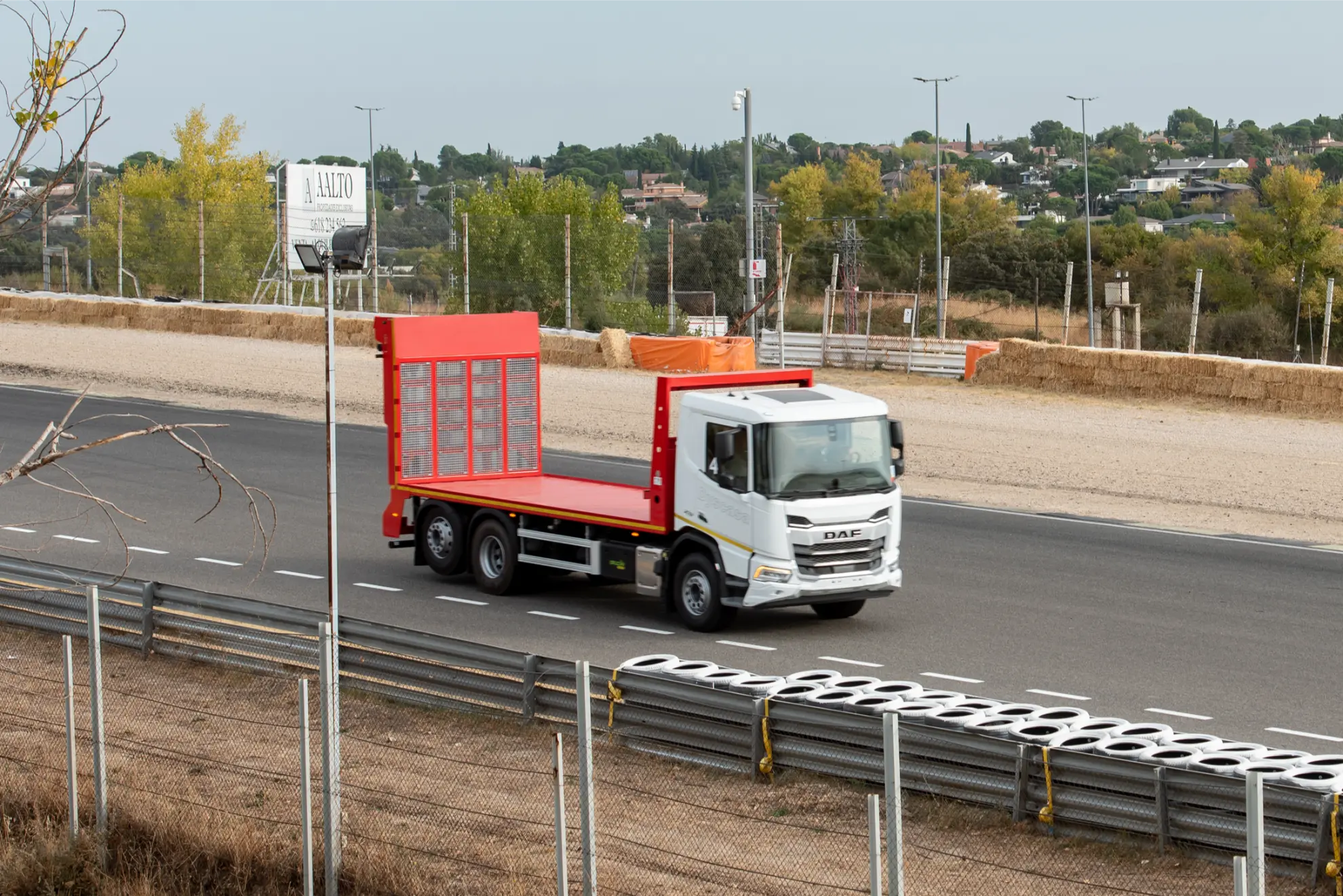 Un camión de plataforma blanco y rojo circula por una carretera cerca de un hipódromo vallado, con una exuberante vegetación de fondo, mientras un cartel de Borrador automático llama la atención.