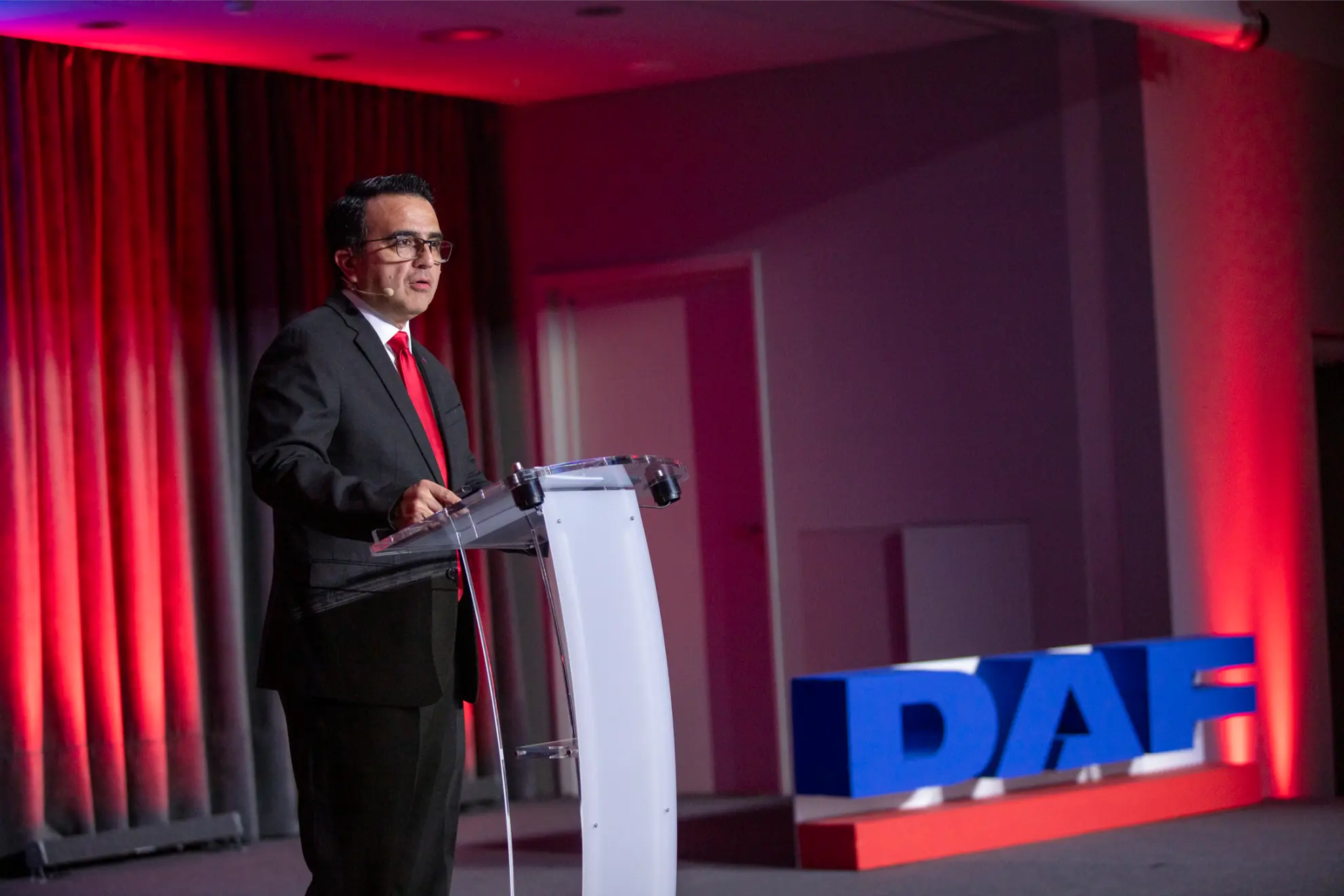 Hombre con traje y corbata roja hablando en un podio, con iluminación roja y grandes letras "DAF" de fondo, presentando detalles sobre el nuevo sistema de borrador automático.