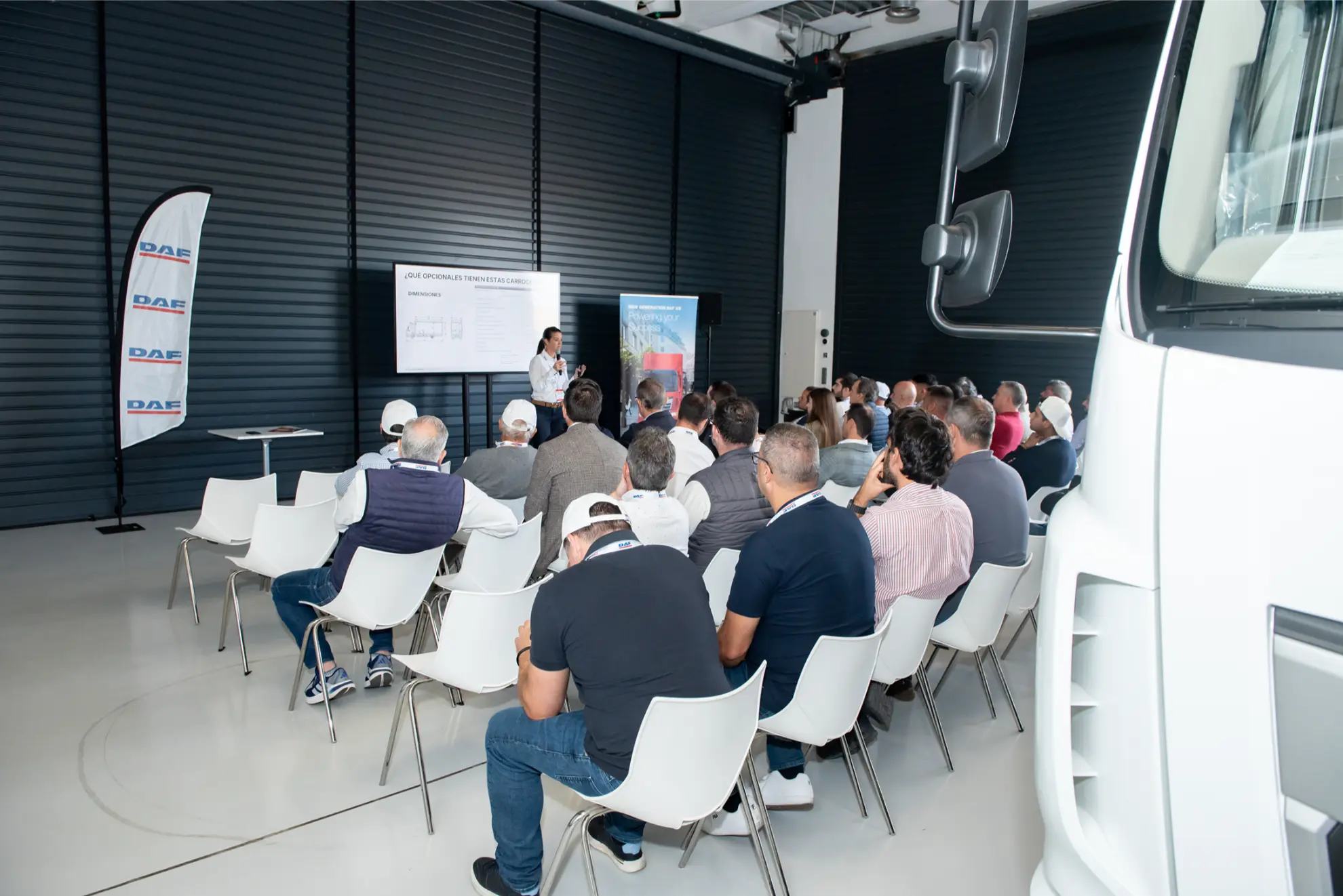 Personas sentadas en sillas blancas asisten a una presentación en una sala luminosa y moderna, con un camión y un borrador automático visibles en primer plano.