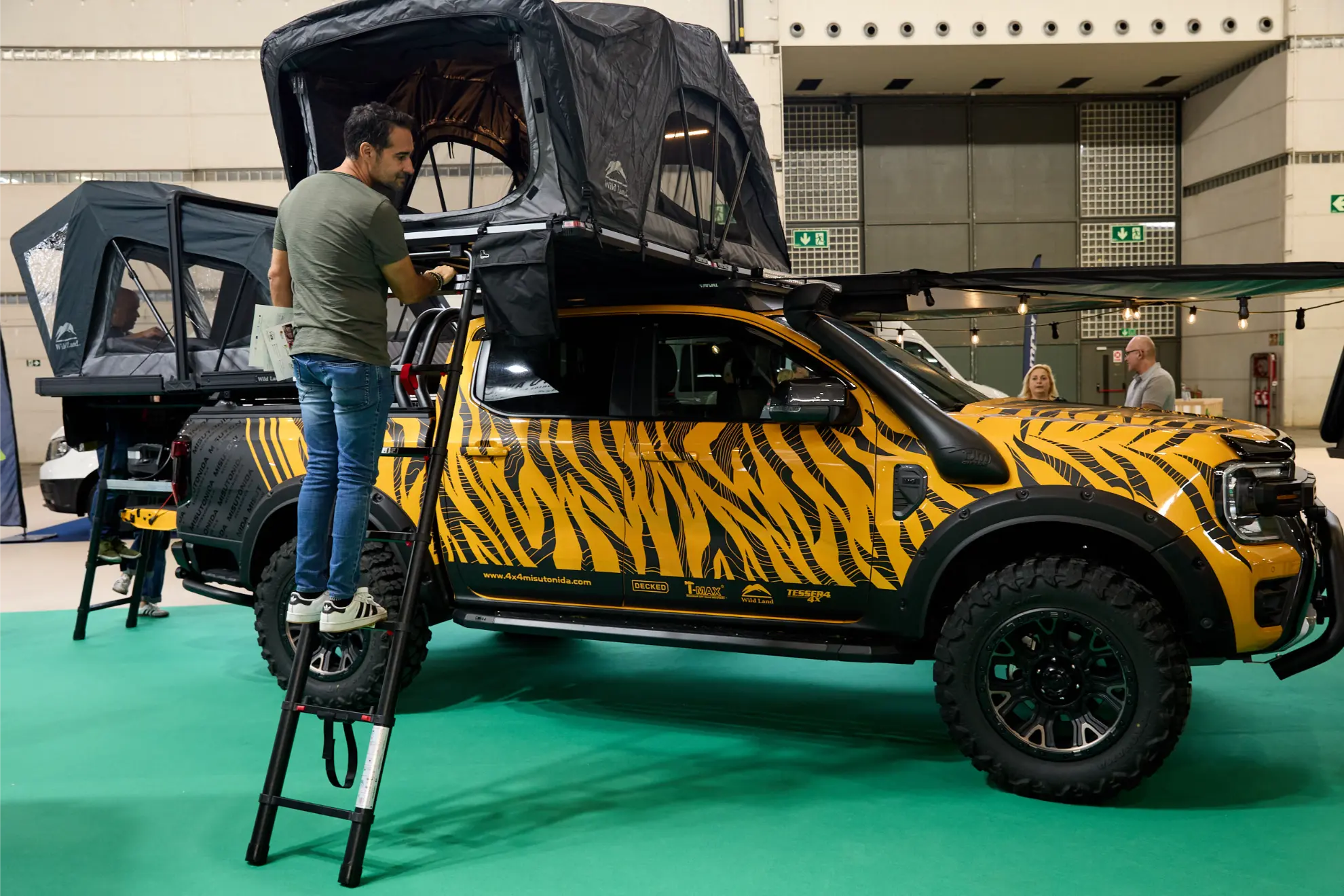 En el Salón del Caravaning, un hombre monta una tienda de campaña en el techo de una furgoneta camper con rayas de tigre utilizando una pequeña escalera.