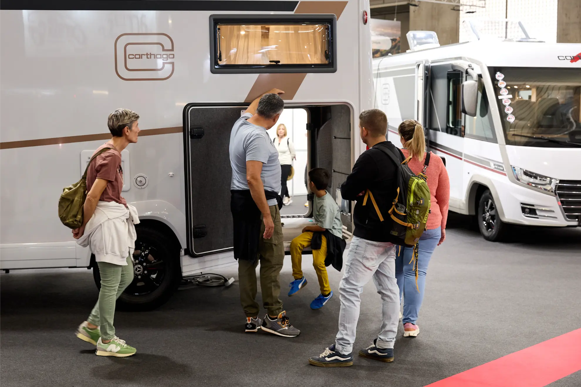 Cinco personas examinan el compartimento de almacenamiento de una autocaravana aparcada en el Salón del Caravaning, una feria de vehículos recreativos de interior.