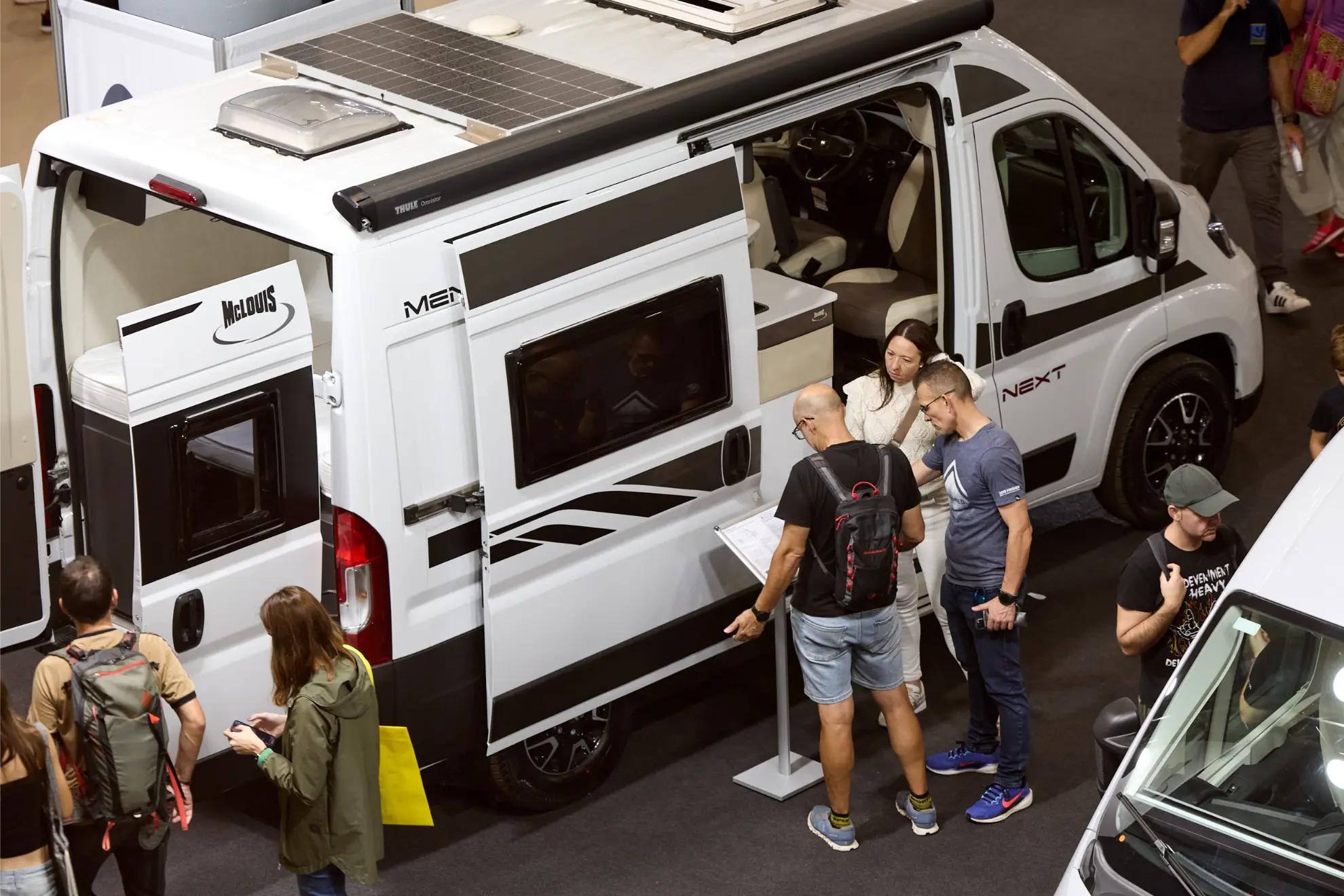 La gente examina una autocaravana blanca con paneles solares en el Salón del Caravaning, leyendo información en un stand y explorando lo último en autocaravanas.