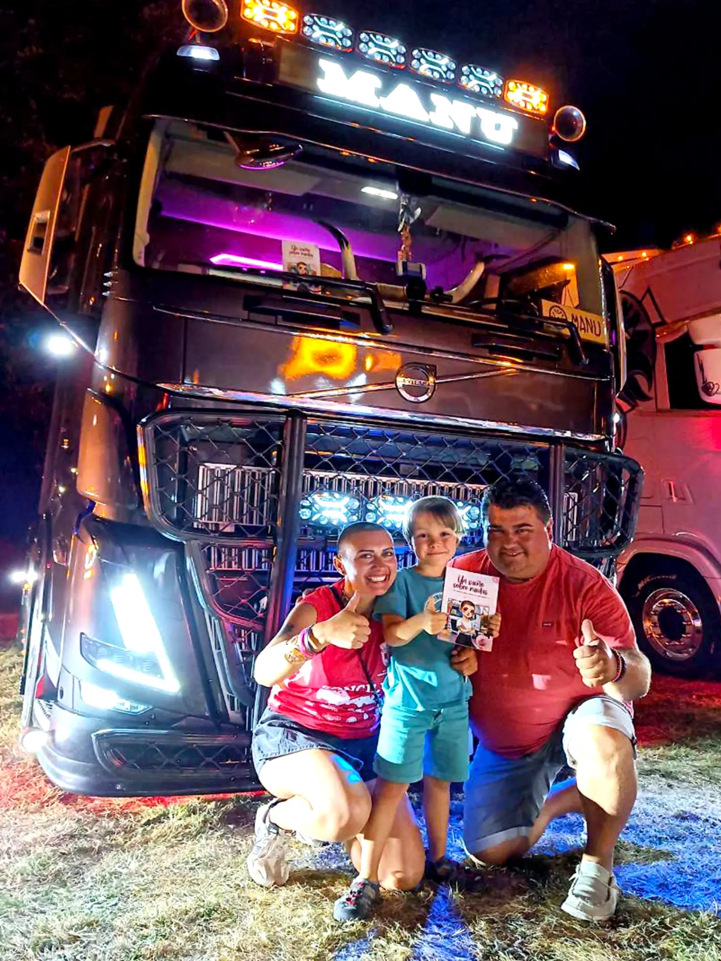 Una familia sonriente irradia alegría ubicua mientras posa de noche frente a una cautivadora camioneta iluminada, dando pulgares arriba.