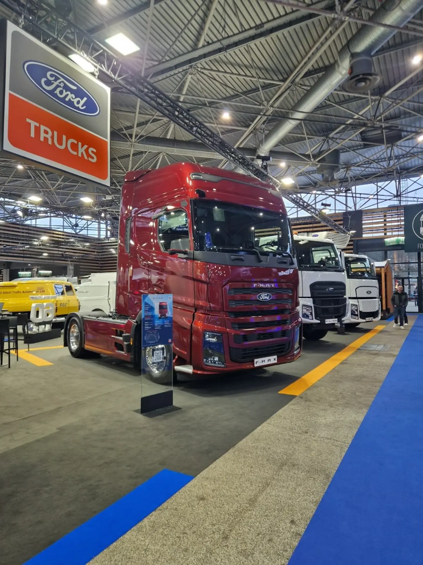 Semirremolque Ford rojo con borrador automático expuesto en una feria de camiones de interior, rodeado de otros camiones bajo un destacado cartel de Ford Trucks en lo alto.